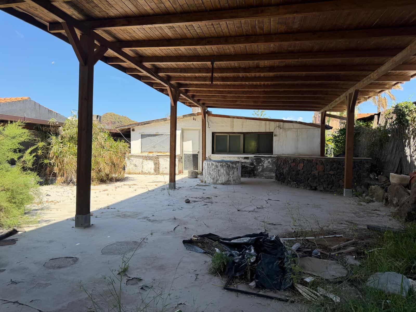 Struttura esterna di una casa abbandonata con tetto in legno e pavimentazione in cemento, ambiente esterno trascurato con detriti e vegetazione selvatica.