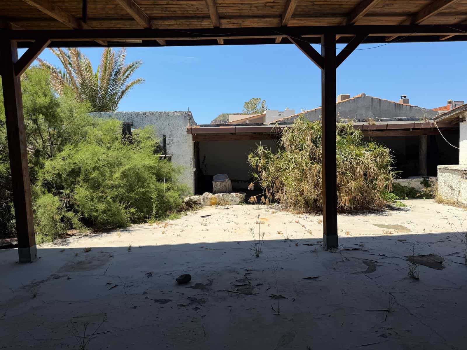 VISTA DALL'ESTERNO DI UN'AREA ABITABILE ABANDONATA, con vegetazione selvaggia e strutture in rovina, sotto un portico in legno, con cielo azzurro e palme sullo sfondo.