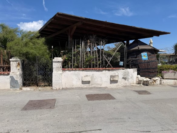 Un'area esterna con struttura in legno e tettoia, circondata da un muro in pietra e recinzione in rete metallica, con piante e vegetazione, sotto un cielo sereno.