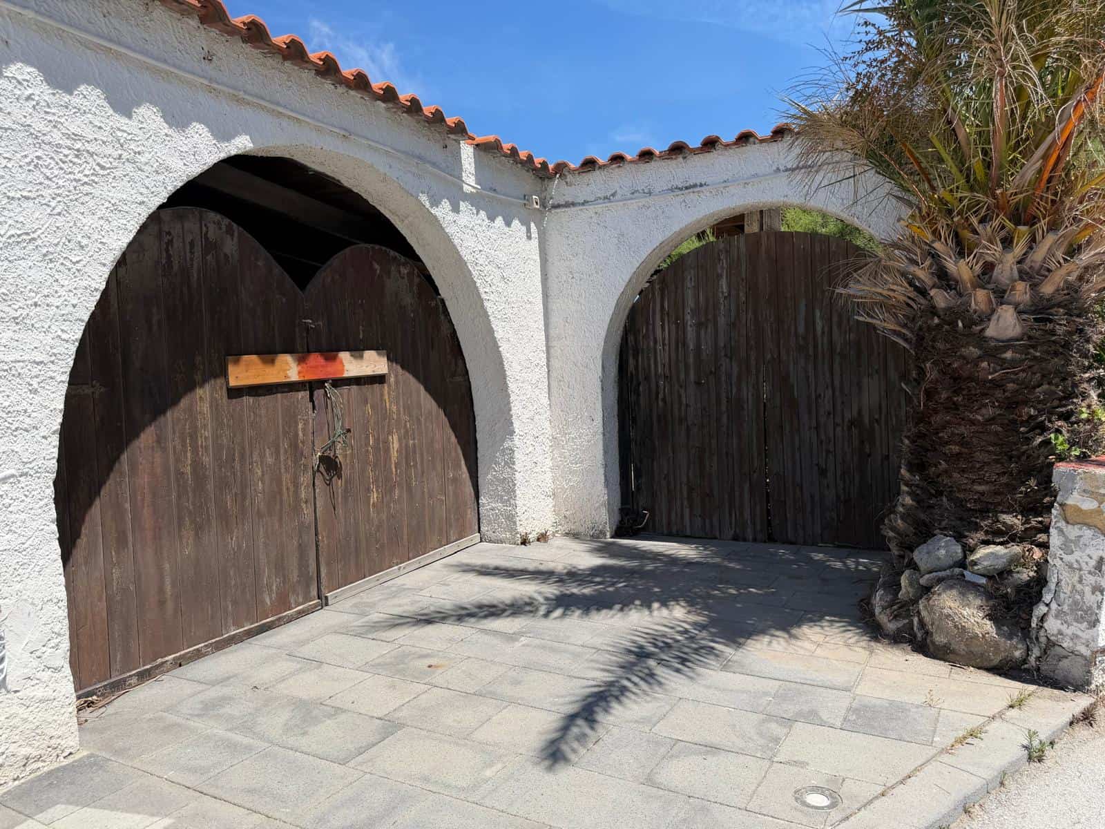 Barriera di ingresso in legno con archi in muratura bianca, circondata da piante tropicali e pietre decorative, sotto un cielo sereno, in un ambiente esterno elegante e accogliente.