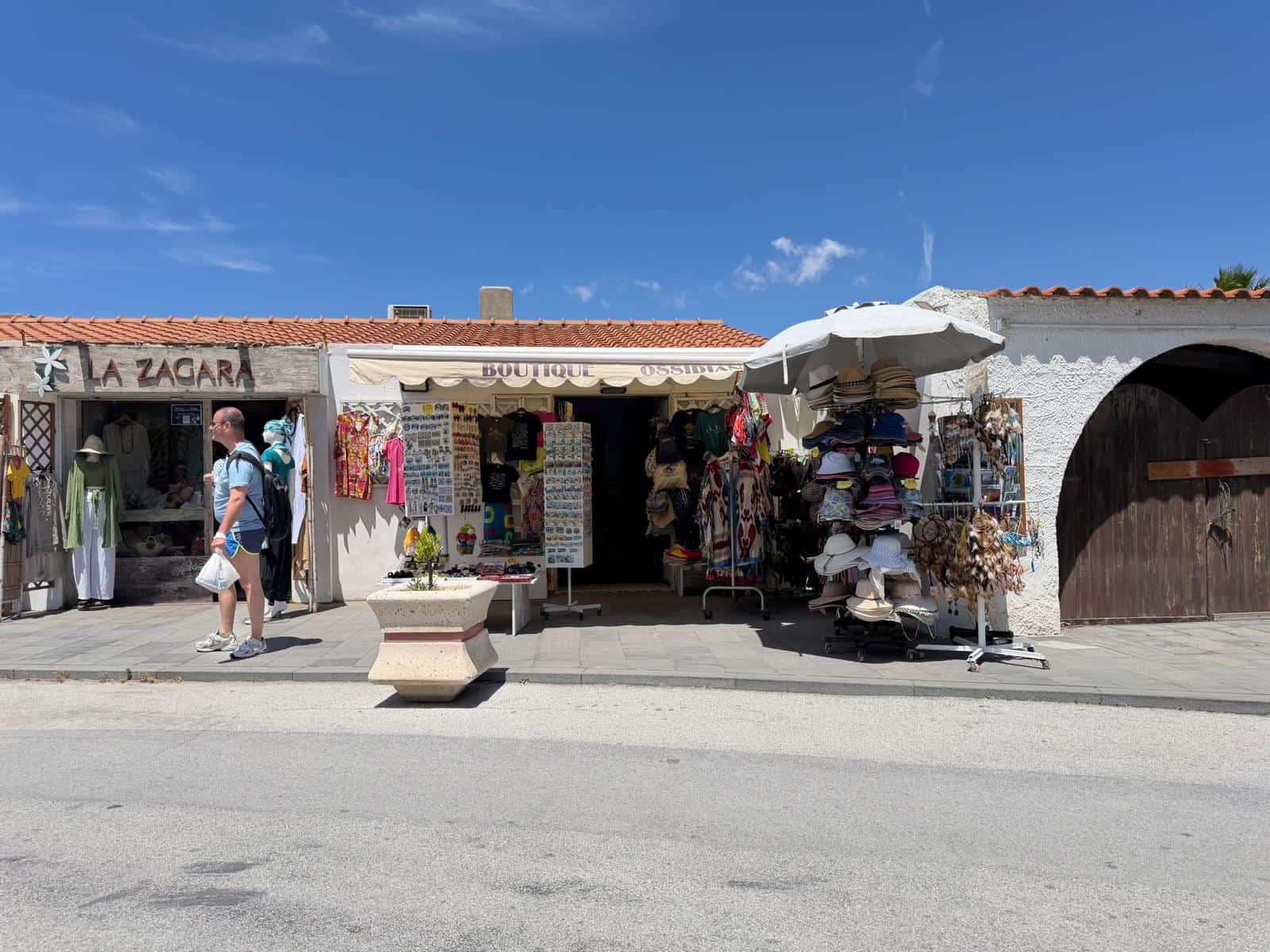 Vendita di souvenir e articoli locali in un negozio all'aperto con espositori di cappelli, abbigliamento e oggetti decorativi, situato in una zona turistica soleggiata e vivace.
