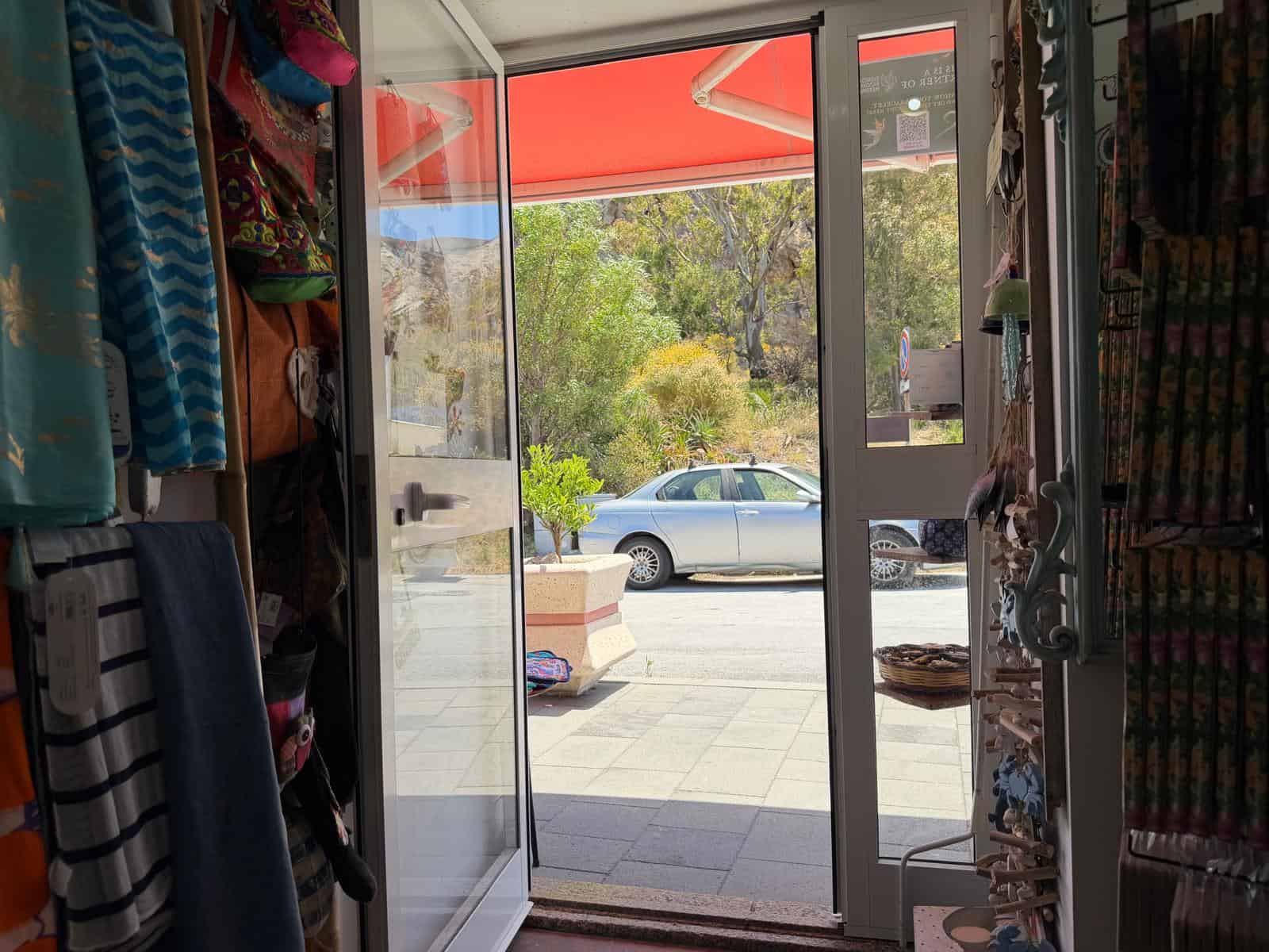 Tappeto e accessori di moda all'ingresso di un negozio di abbigliamento con vetrina e vista sulla strada, con auto parcheggiate e vegetazione naturale sullo sfondo.