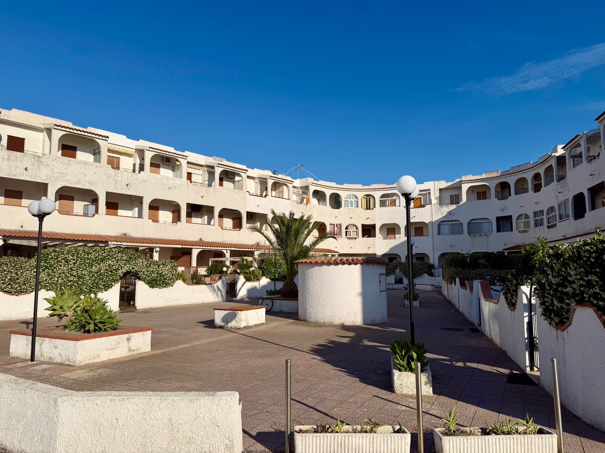 Complesso residenziale con edificio bianco a più piani, cortile centrale con piante e lampioni, cielo sereno, stile mediterraneo, NextCasa.