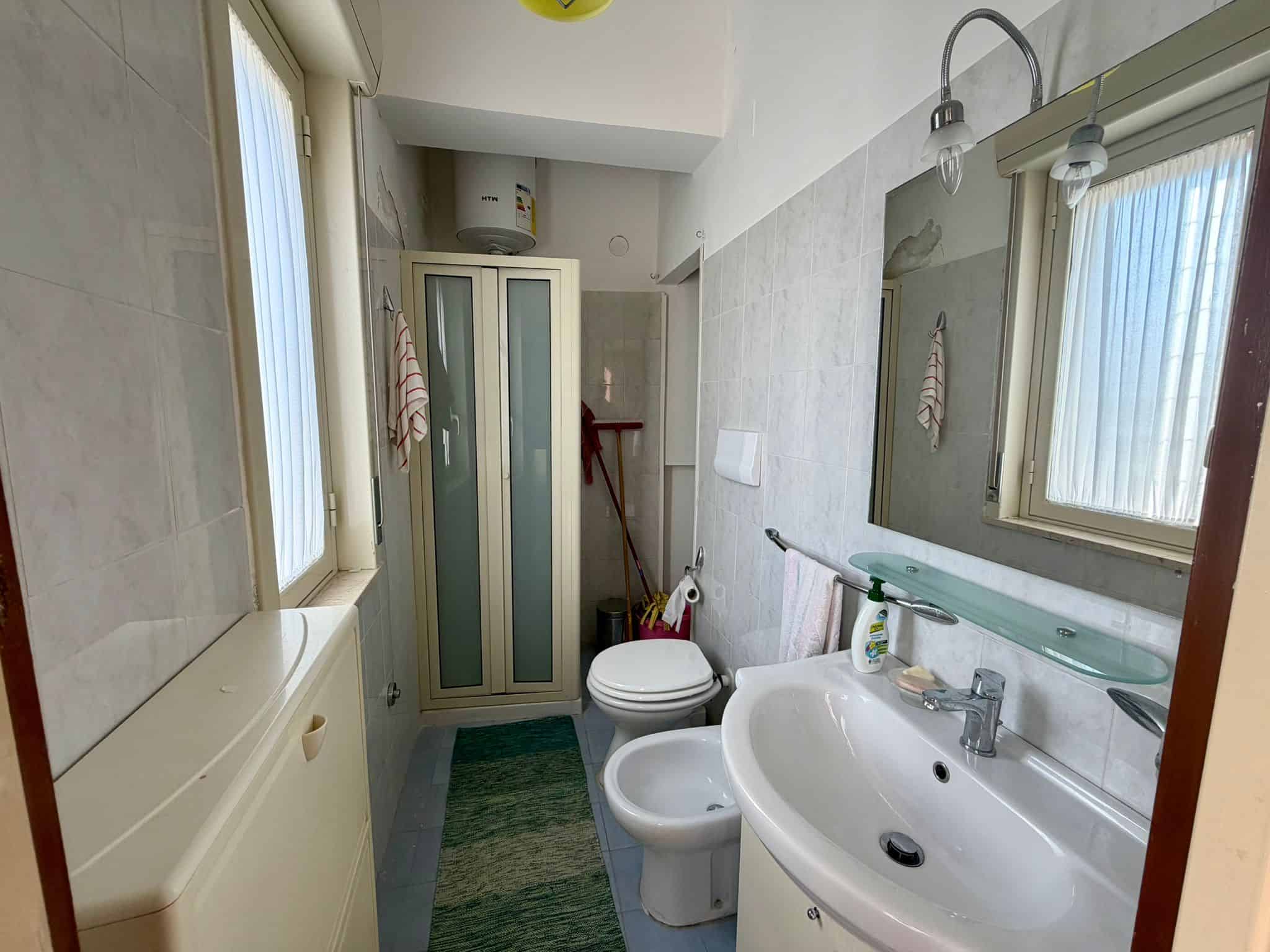 Lavabo e servizi igienici in un bagno di casa con pareti rivestite in piastrelle chiare, grande specchio sopra il lavandino, finestra per l'illuminazione naturale, e accessori essenziali come dispense.