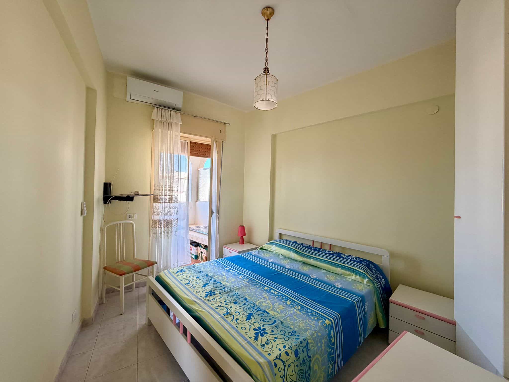 Camera da letto luminosa con pareti gialle, letto matrimoniale con copriletto blu e finestra con tende trasparenti, ambiente accogliente e ben illuminato.