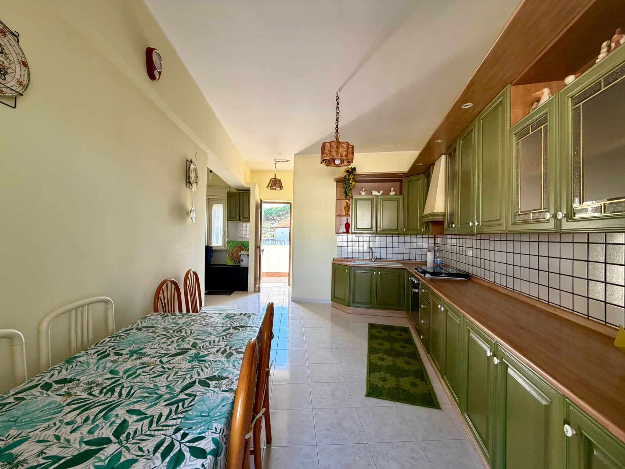 Cucina spaziosa con mobili verdi, tavolo con tovaglia a foglie e illuminazione calda, ambiente luminoso e accogliente in stile rustico.