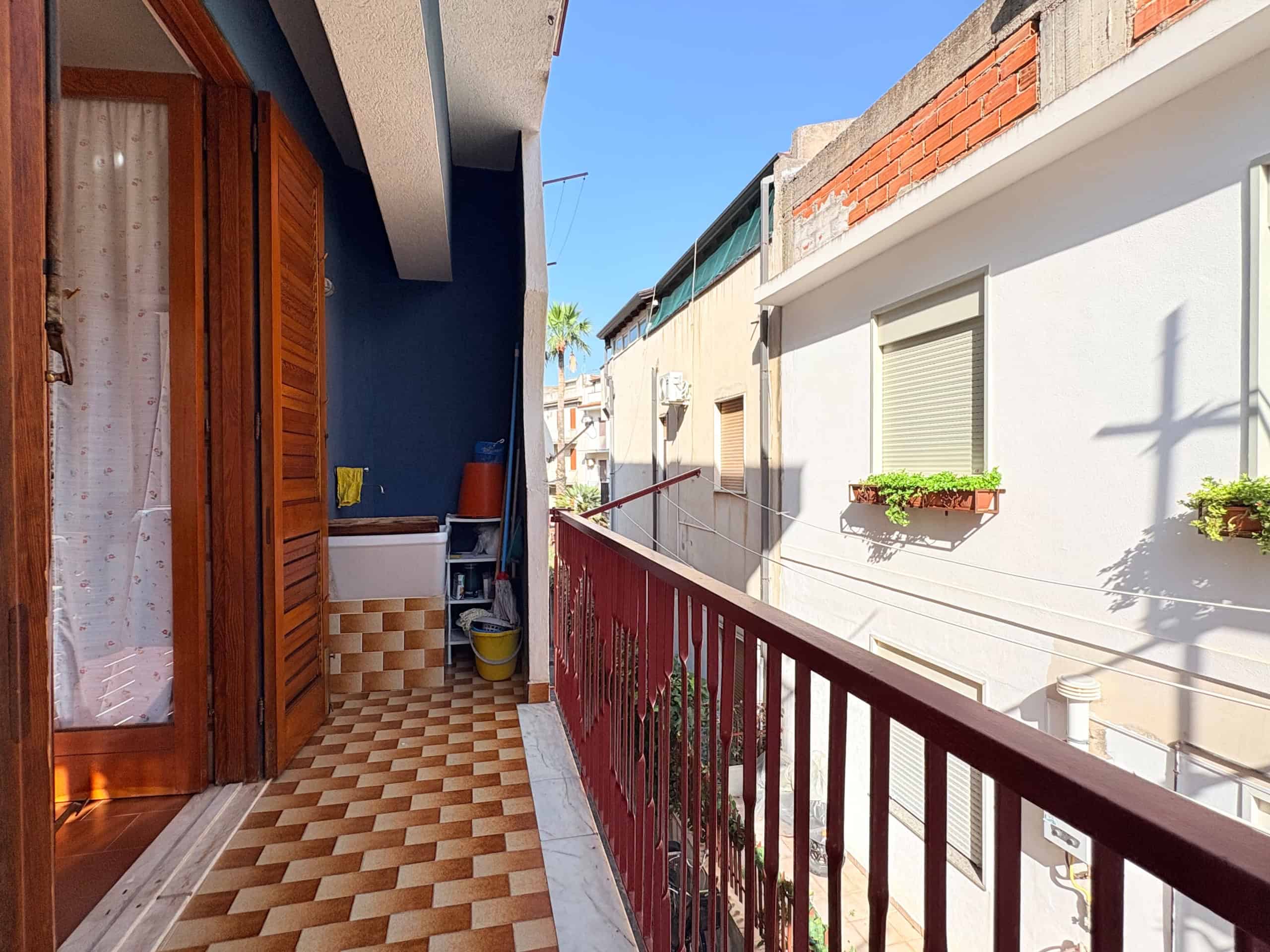 Balcone di un appartamento urbano con ringhiera rossa, vista su edifici residenziali con piante in vaso, cielo sereno e luce naturale, ideale per relax e vita all'aperto.