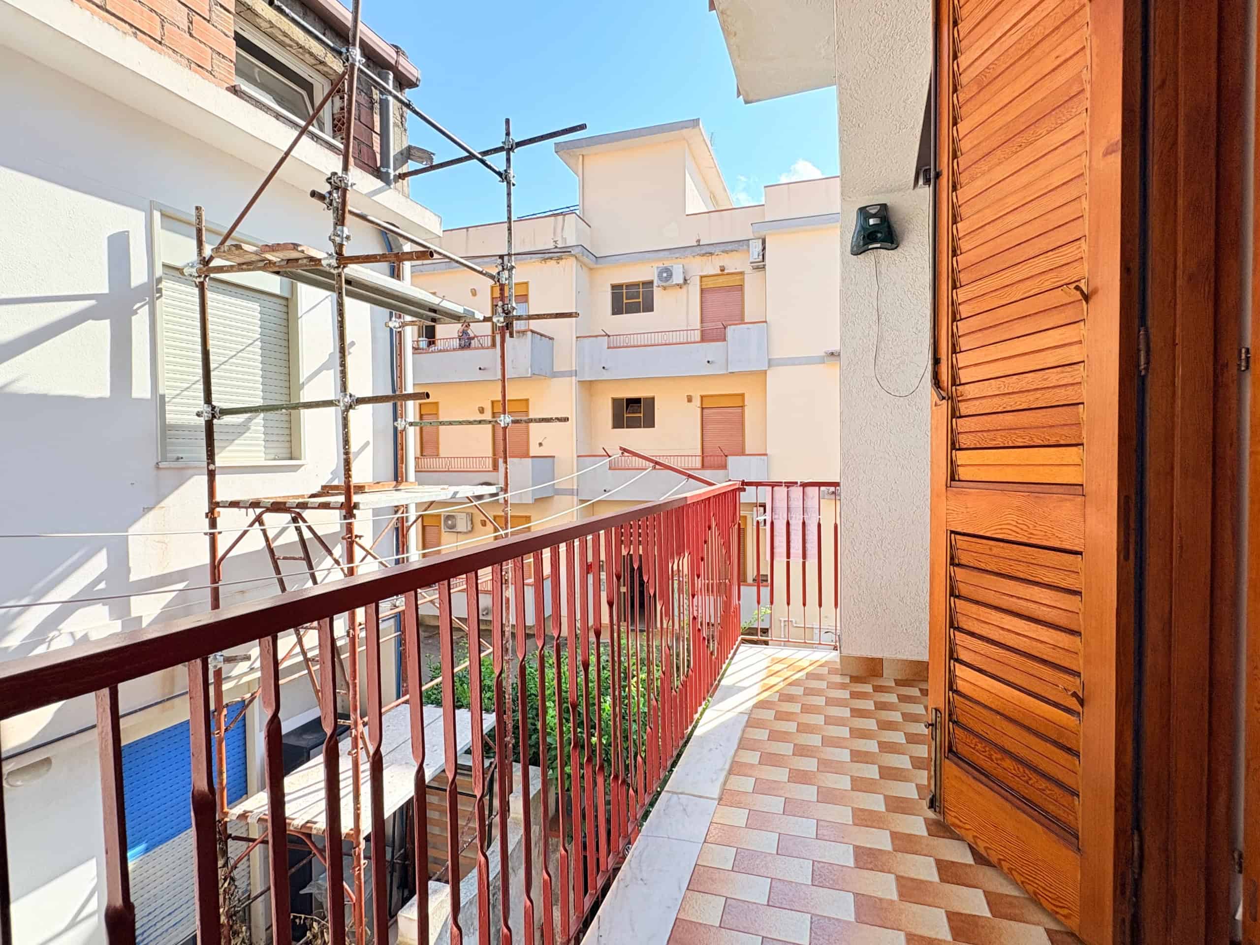 Balcone di un appartamento in fase di ristrutturazione, con ringhiera rossa e pavimento in piastrelle beige e marroni, vista su edifici circostanti e cielo azzurro, con scaffali di legno e pareti bian.