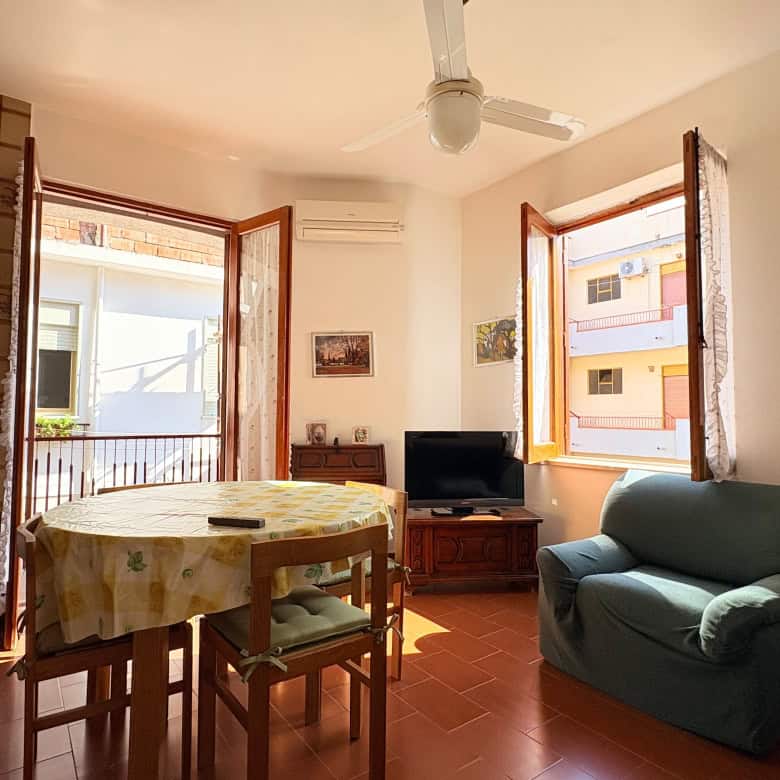 Ampia zona giorno con due finestre aperte, illuminata da luce naturale, arredi accoglienti e stile semplice, ideale per vivere momenti di relax e convivialità in un appartamento a NextCasa.
