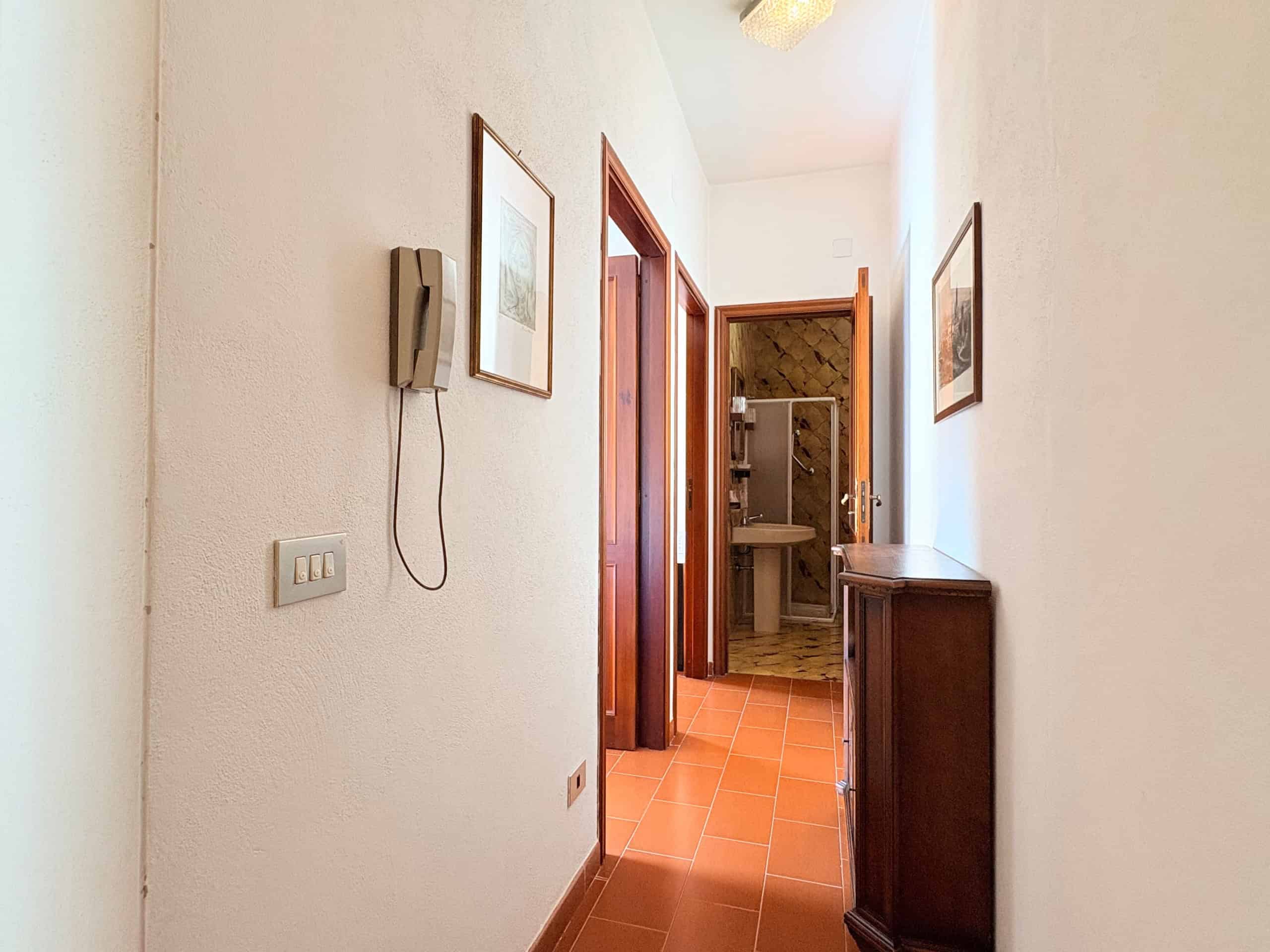 Elegante corridoio interno di casa con pareti bianche, arredi in legno e accessori decorativi, che conduce a un bagno ben illuminato e accogliente, ideale per ambienti residenziali di qualità.