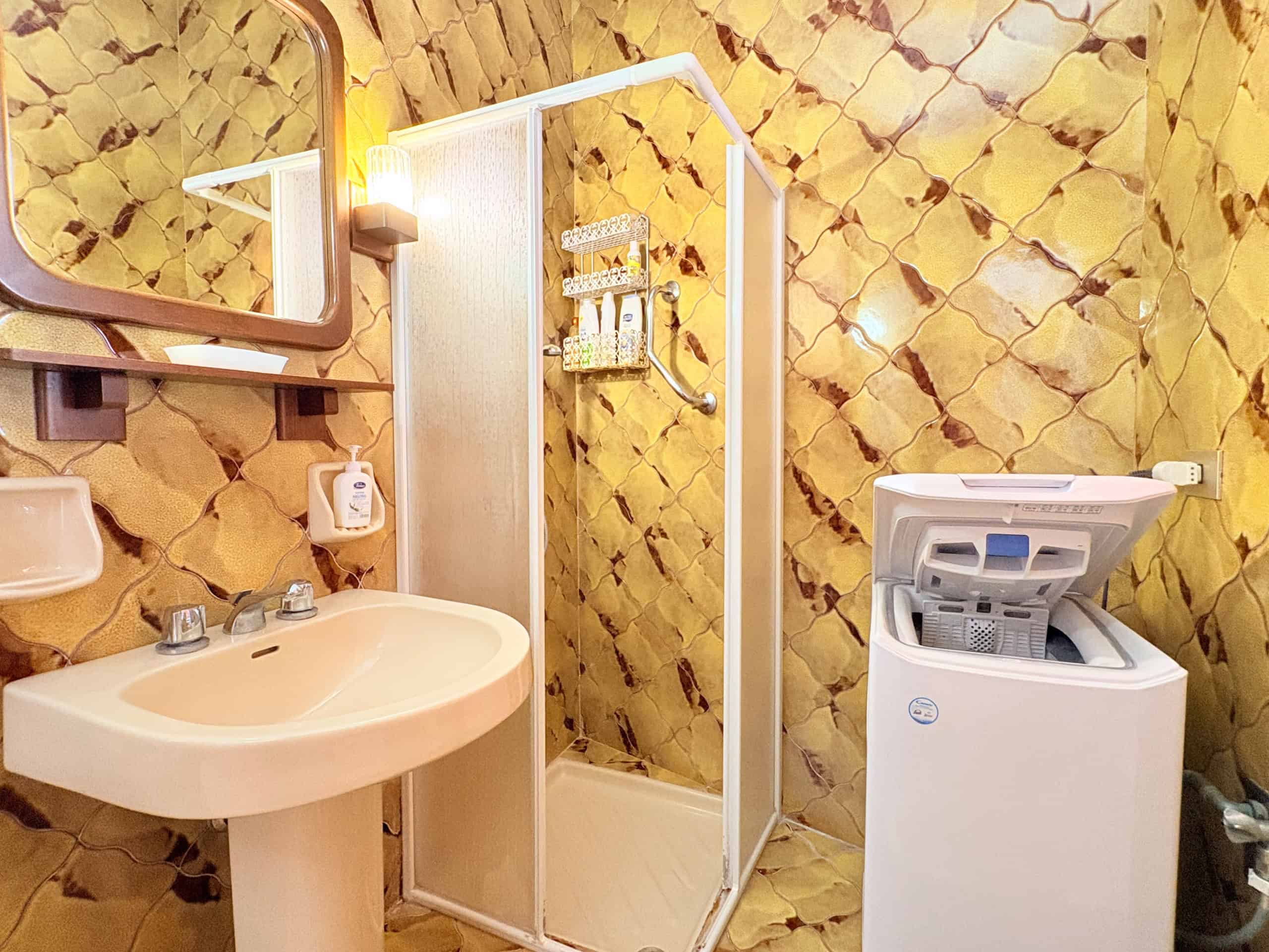 Lavabo e doccia in un bagno con pareti rivestite in piastrelle gialle a mosaico, con lavatrice moderna e specchio con luce integrata, ambiente funzionale e ben illuminato.