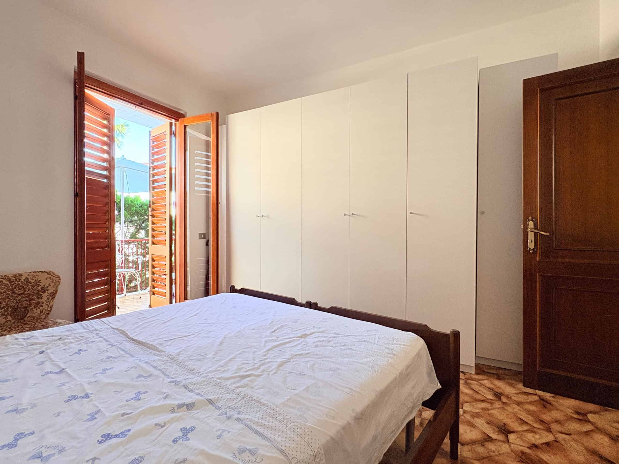 Il letto matrimoniale in una camera da letto luminosa con armadi bianchi e porta finestra che dà su un balcone soleggiato, ideale per case o appartamenti in vendita o affitto.