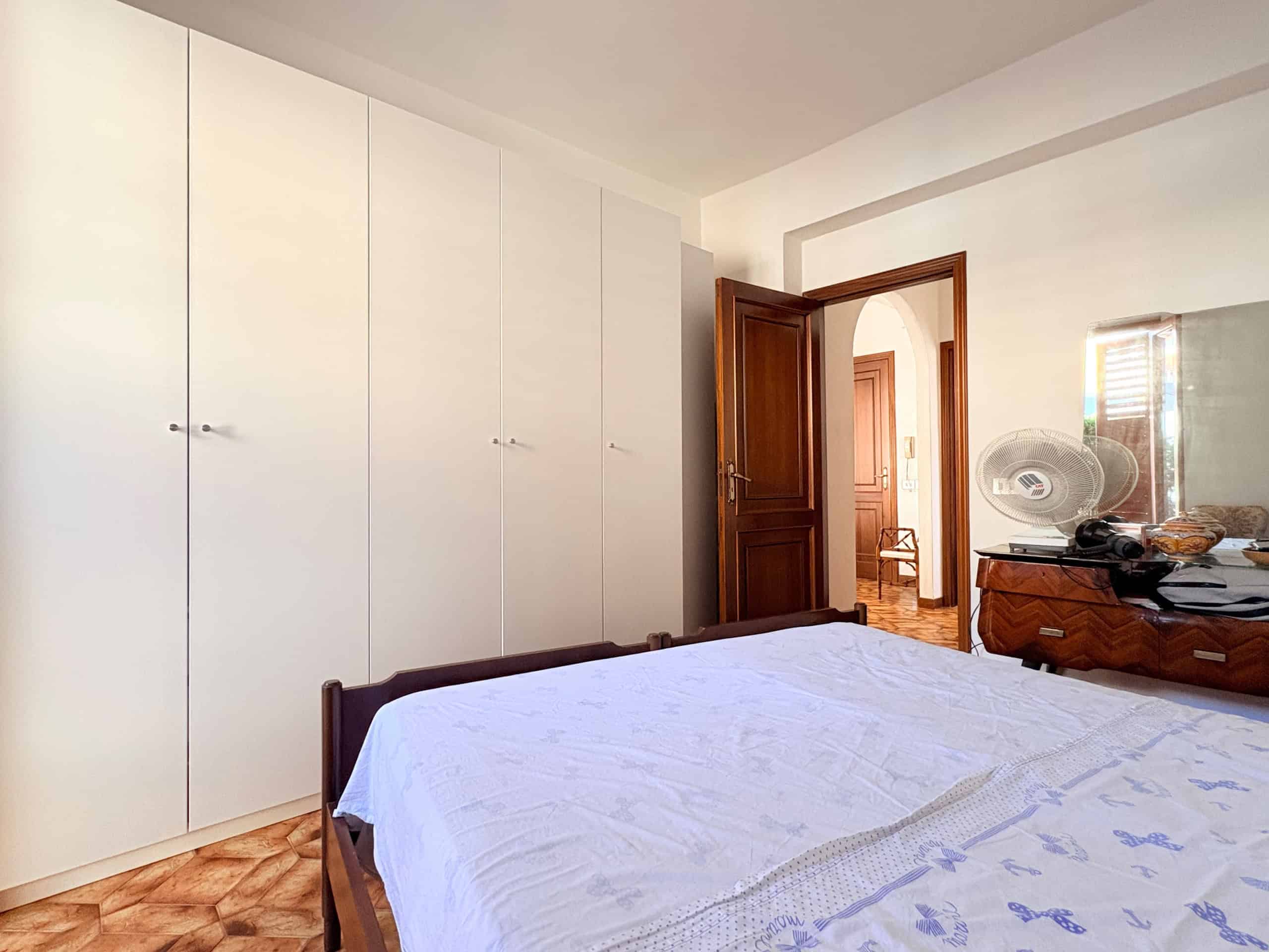 Il letto matrimoniale in una camera da letto luminosa con armadi a muro bianchi e un mobile in legno, con un ventilatore e uno specchio, in un ambiente accogliente e ben organizzato.
