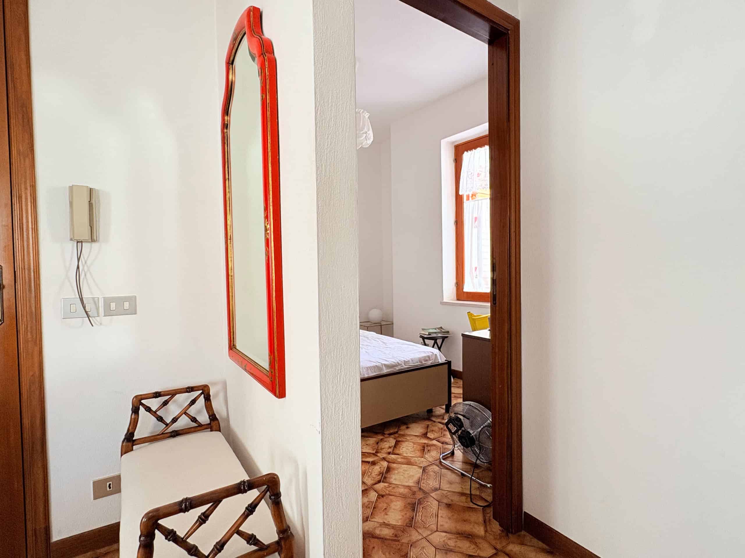 Specchio con cornice rossa in ingresso, sedia in legno e vista sulla camera da letto con finestra luminosa, pavimento in piastrelle di ceramica, ambiente accogliente e luminoso.