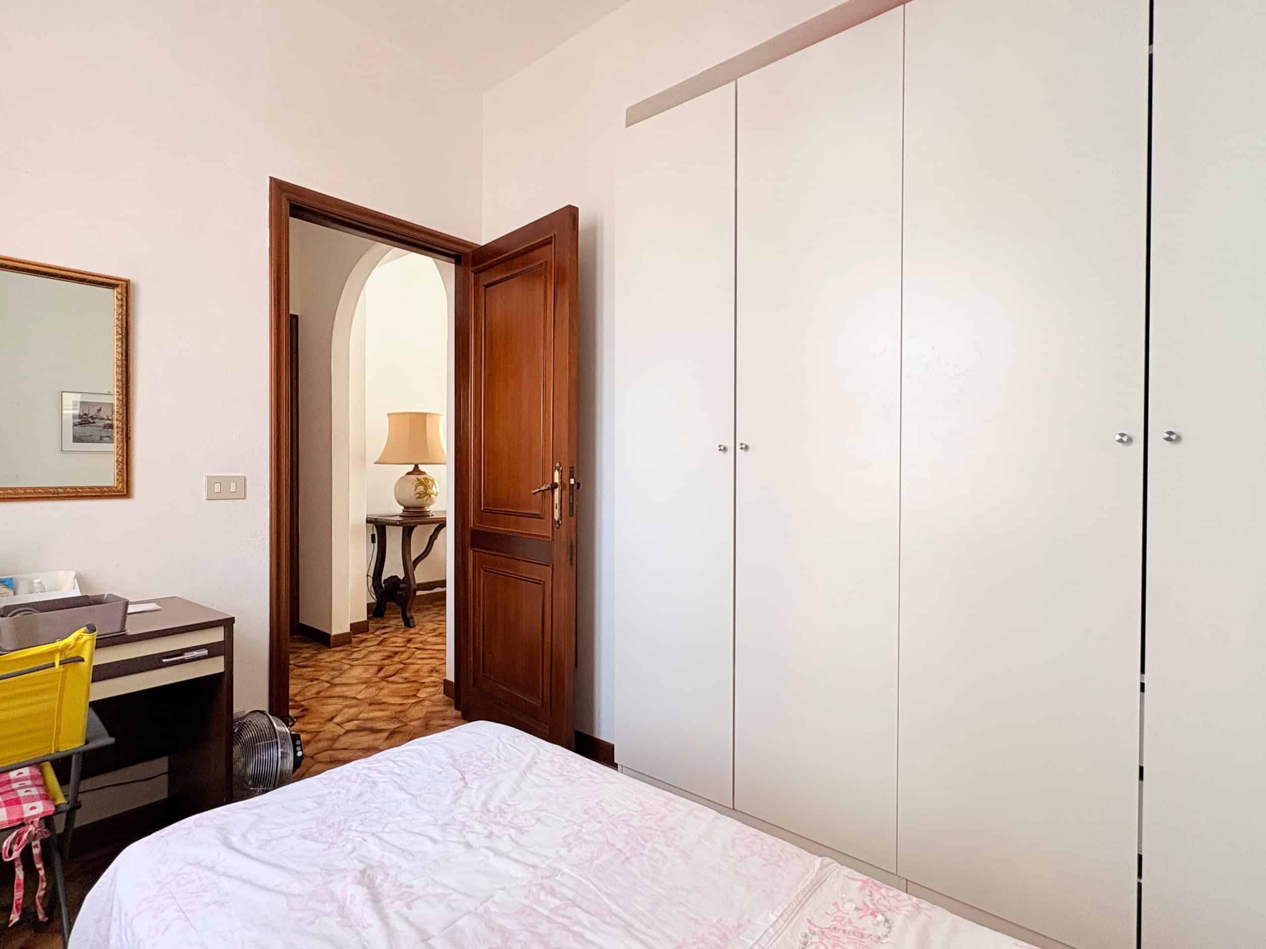 Armadio a muro bianco in una camera da letto con porta in legno aperta, visibile un'area con un tavolo e una lampada in un'altra stanza.