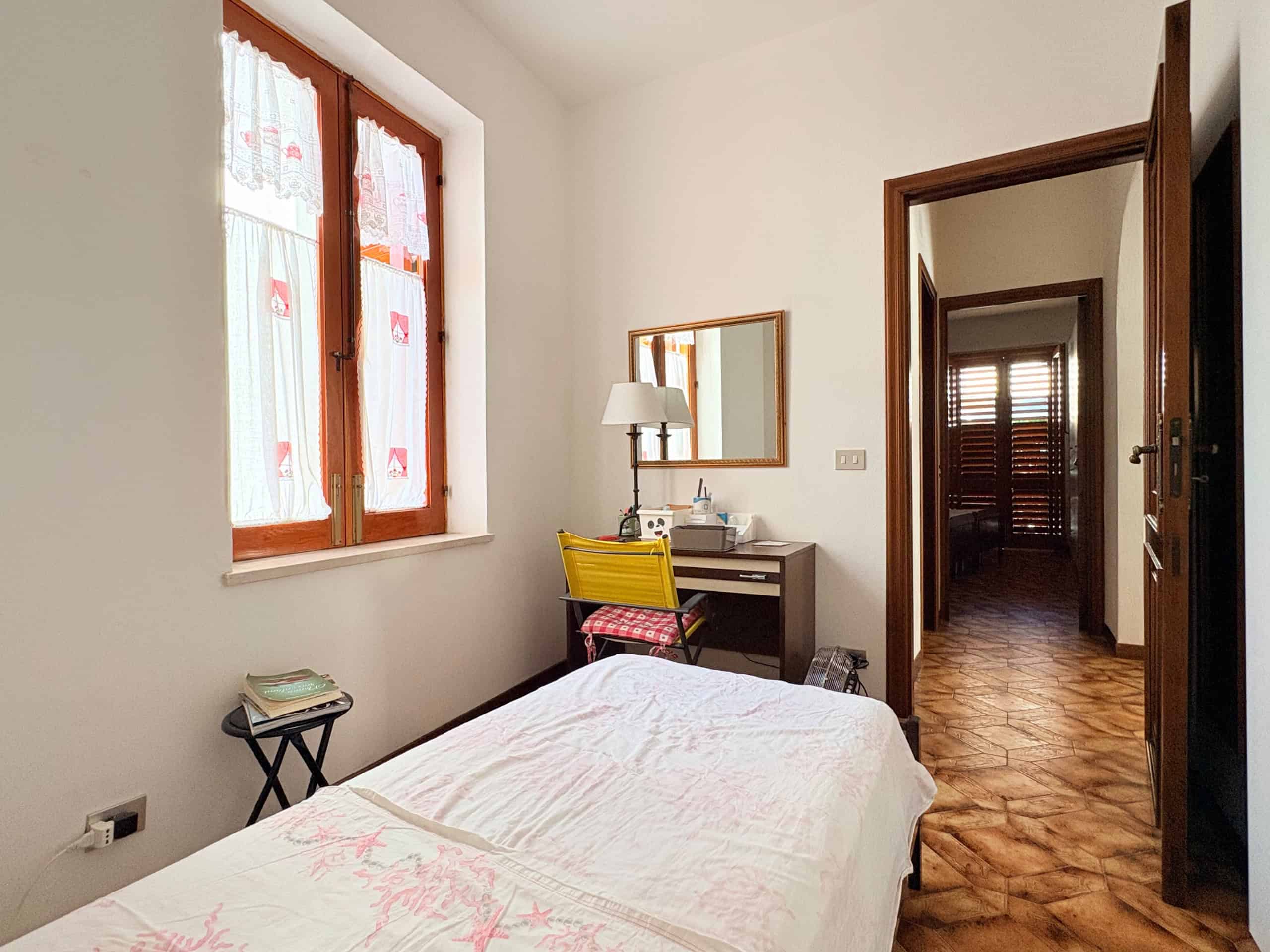 Letto singolo in una camera luminosa con finestra decorata, scrivania con lampada e sedia gialla, pavimento in parquet, ambiente accogliente e ben arredato, ideale per soggiorni confortevoli.