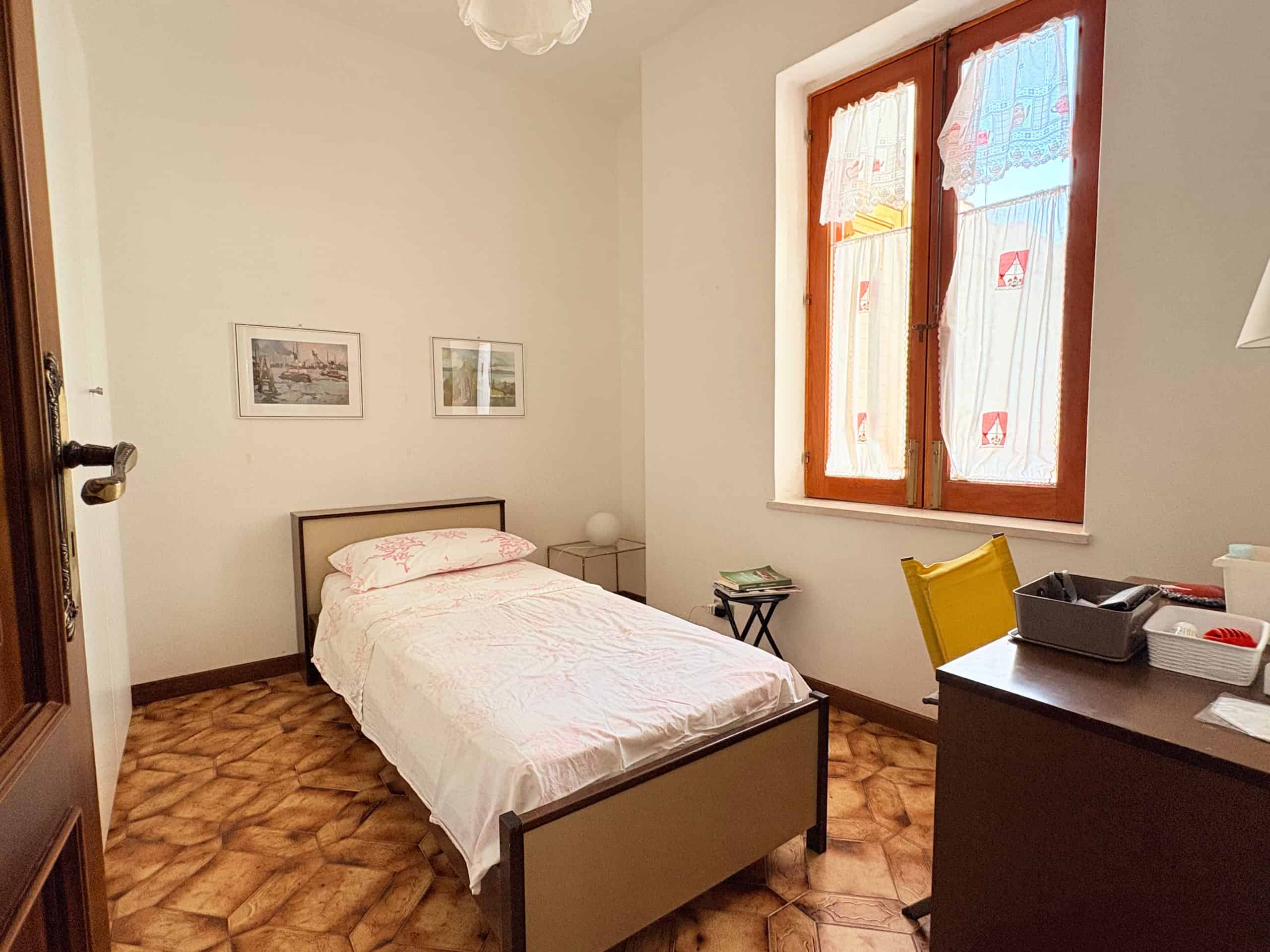 Una camera da letto luminosa con pareti bianche, un letto singolo con copriletto rosa, una finestra grande con tende colorate e un pavimento in parquet, ideale per affitti residenziali o affitti brevi.