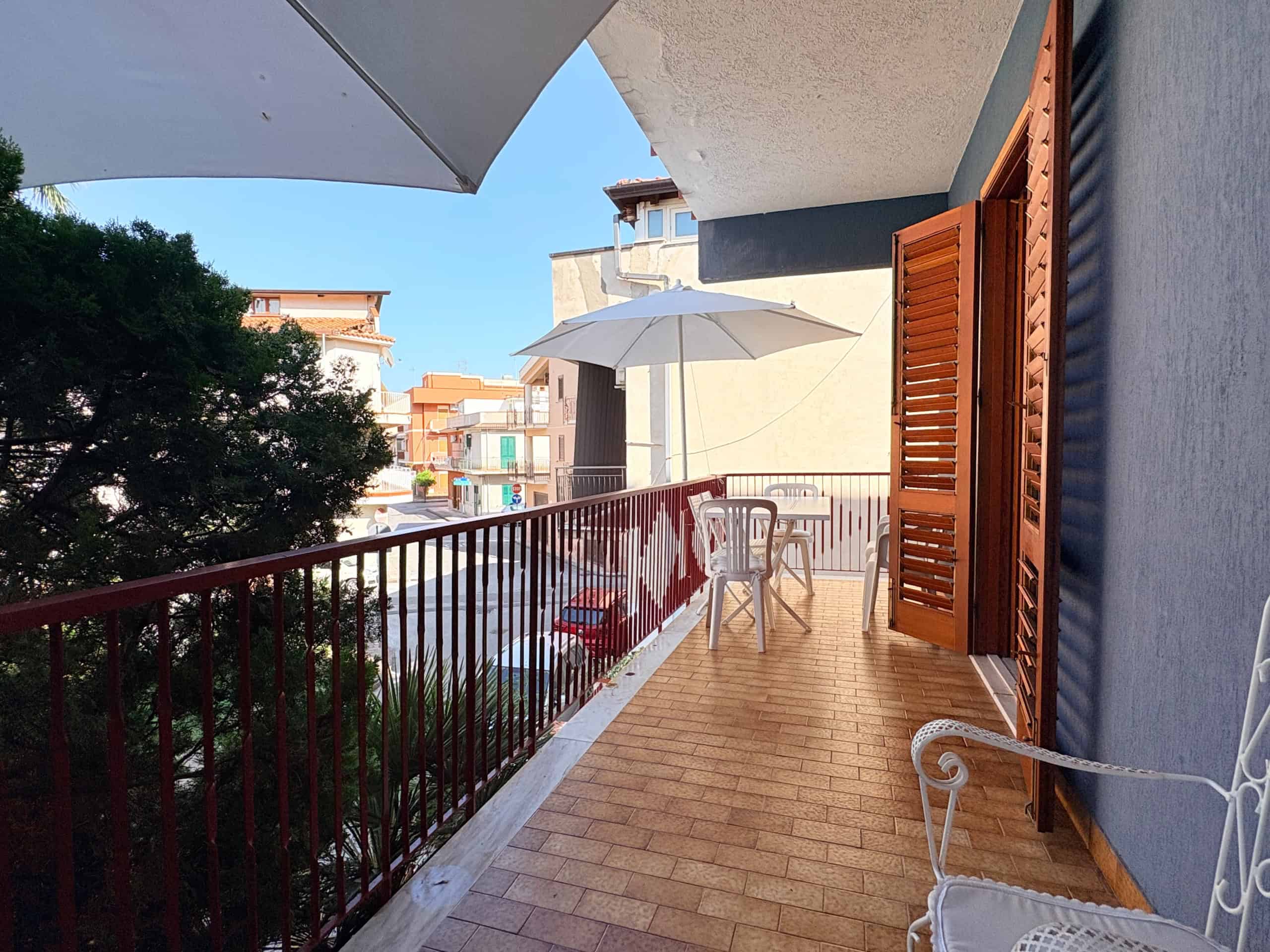 Balcone di un appartamento con vista sulla strada, dotato di ombrellone, sedie e piante, in un quartiere residenziale soleggiato.
