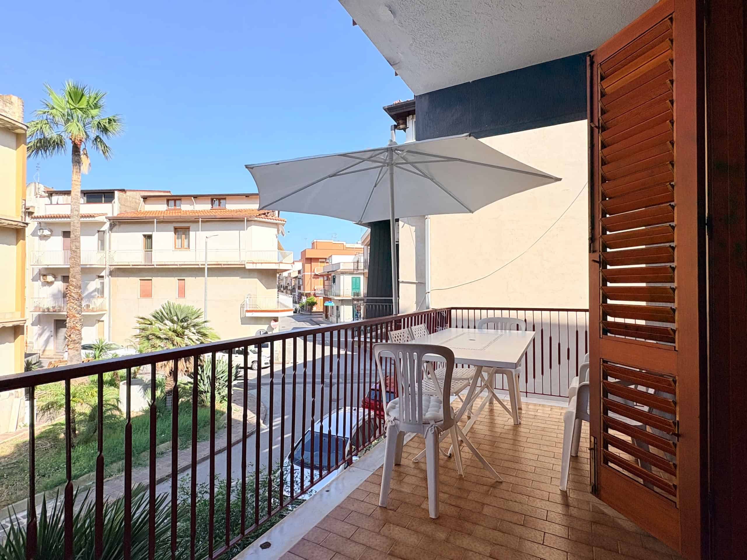 Balcone con tavolo, sedie e ombrellone in un complesso residenziale soleggiato, vista su palazzi e palme, ideale per relax e convivialità.