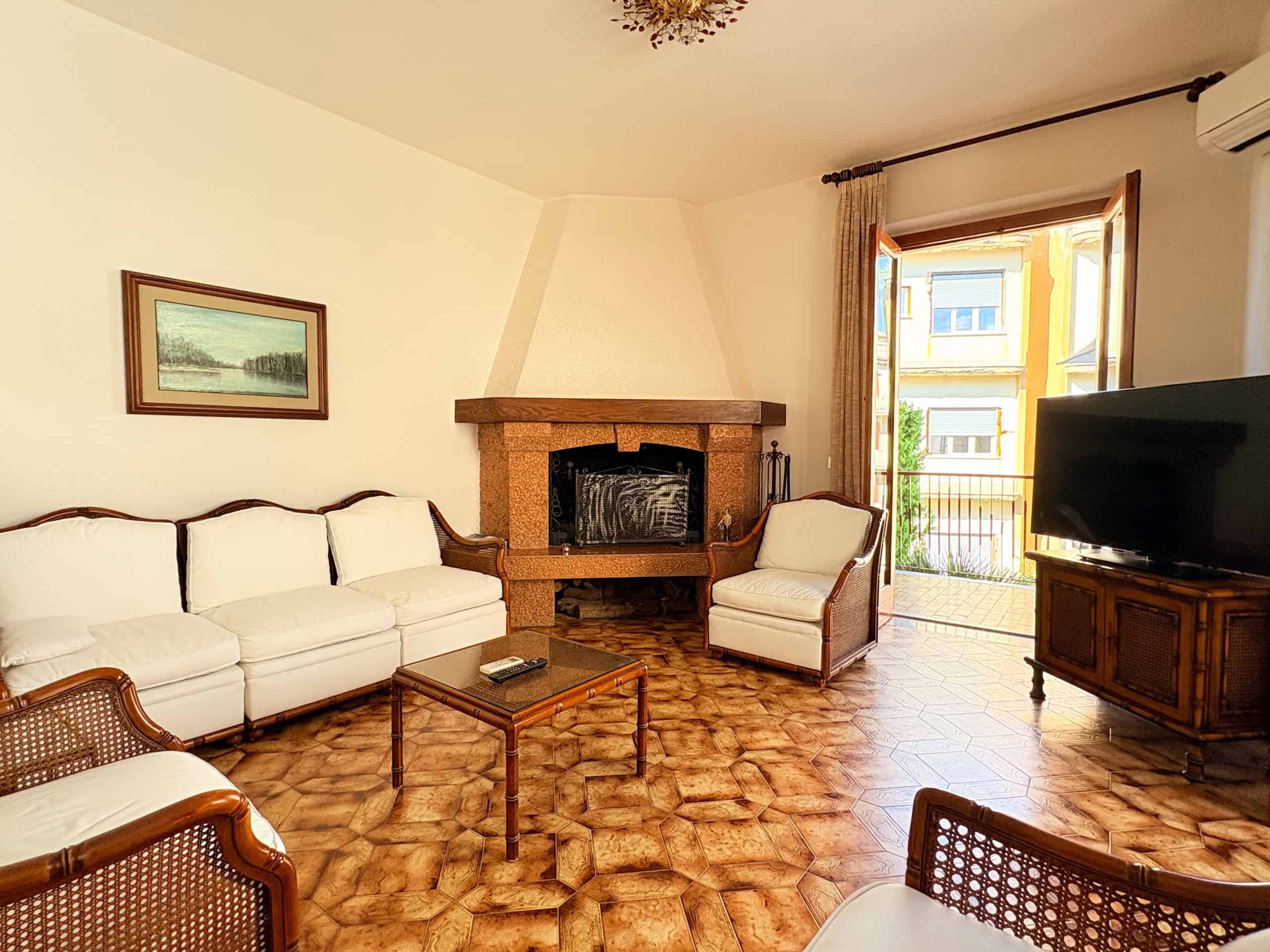 Ampia zona living con divano bianco, camino in pietra, poltrona e televisore, ambiente luminoso con accesso a balcone, arredamento classico, pavimento in parquet, ideale per soggiorno confortevole e a.