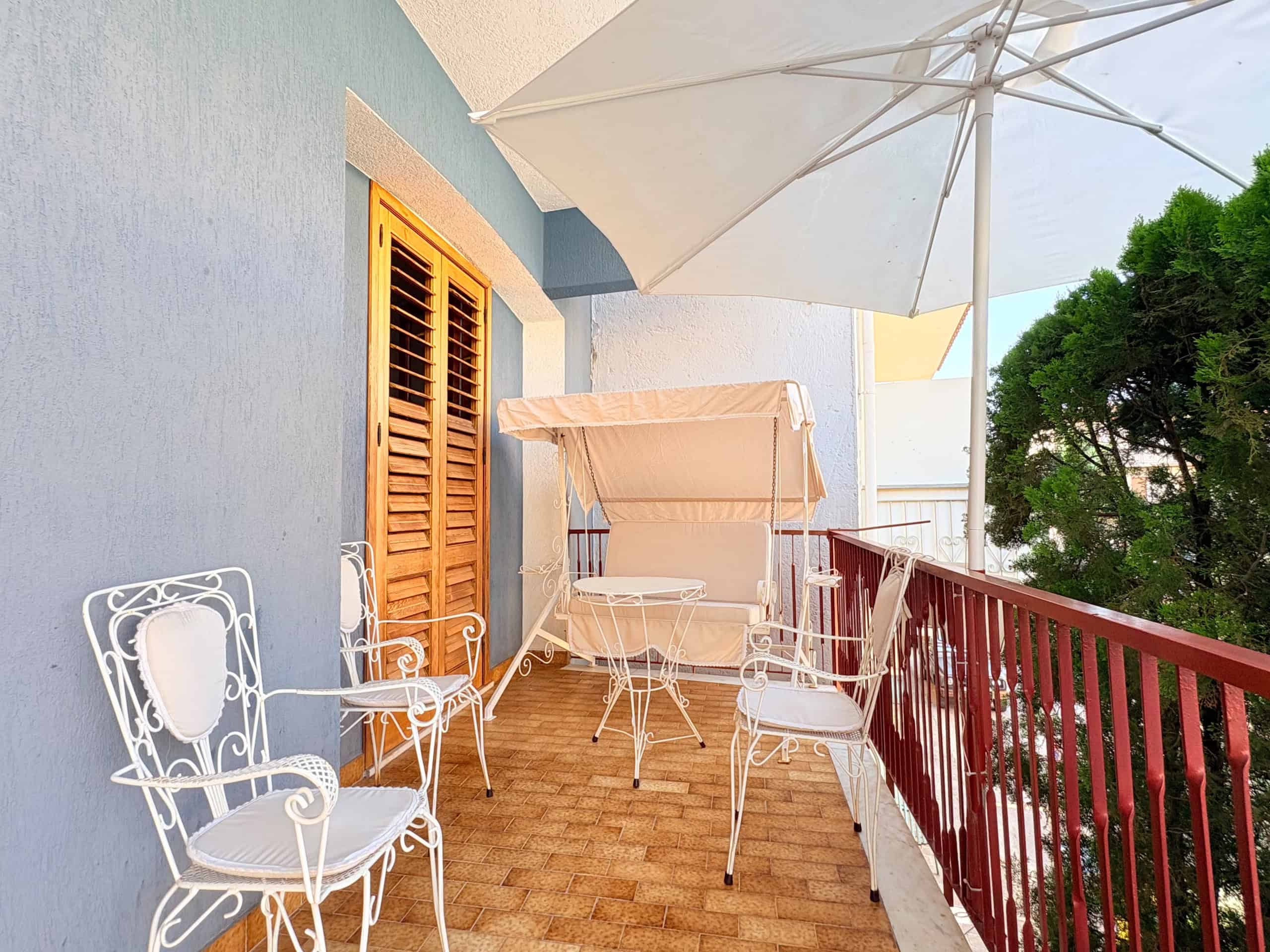 Balcone con sedie in ferro battuto, tavolo e ombrellone, vista su alberi verdi, ambiente esterno luminoso e accogliente, ideale per relax e convivialità all'aperto.
