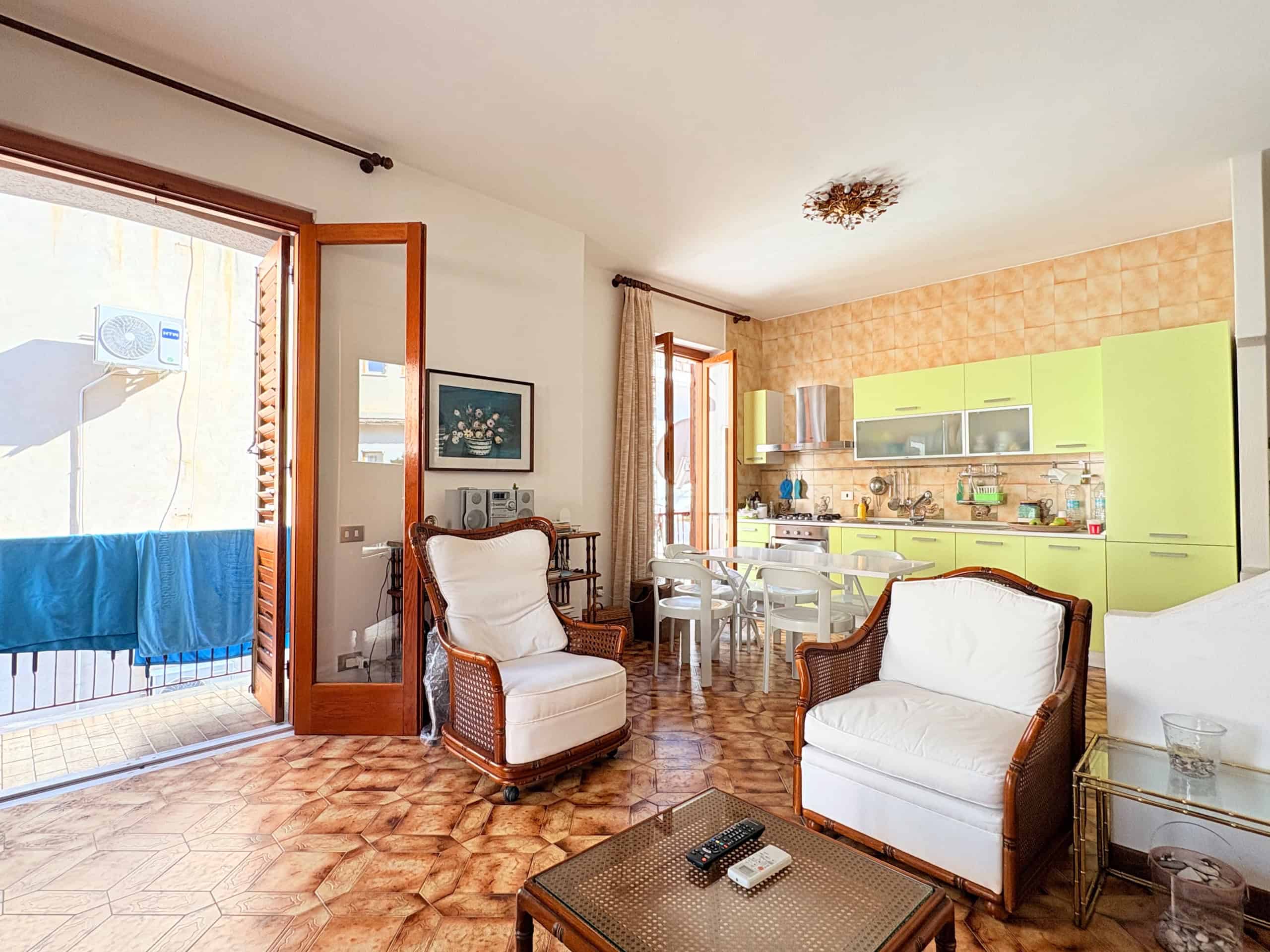 Interno di un soggiorno luminoso con mobili in stile classico e cucina moderna con ante verde lime, balcone con stendibiancheria, ideale per appartamenti in vendita o affitto a NextCasa.