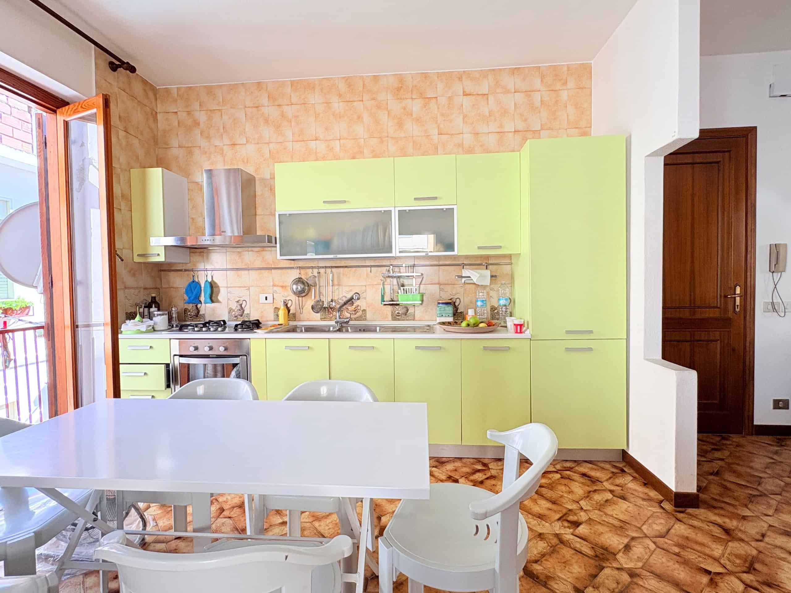 Ampia cucina moderna con mobili verde lime, piano di lavoro in acciaio inox e elettrodomestici, ambiente luminoso con accesso al balcone, pavimento in piastrelle marroni, ideale per la vita quotidiana.