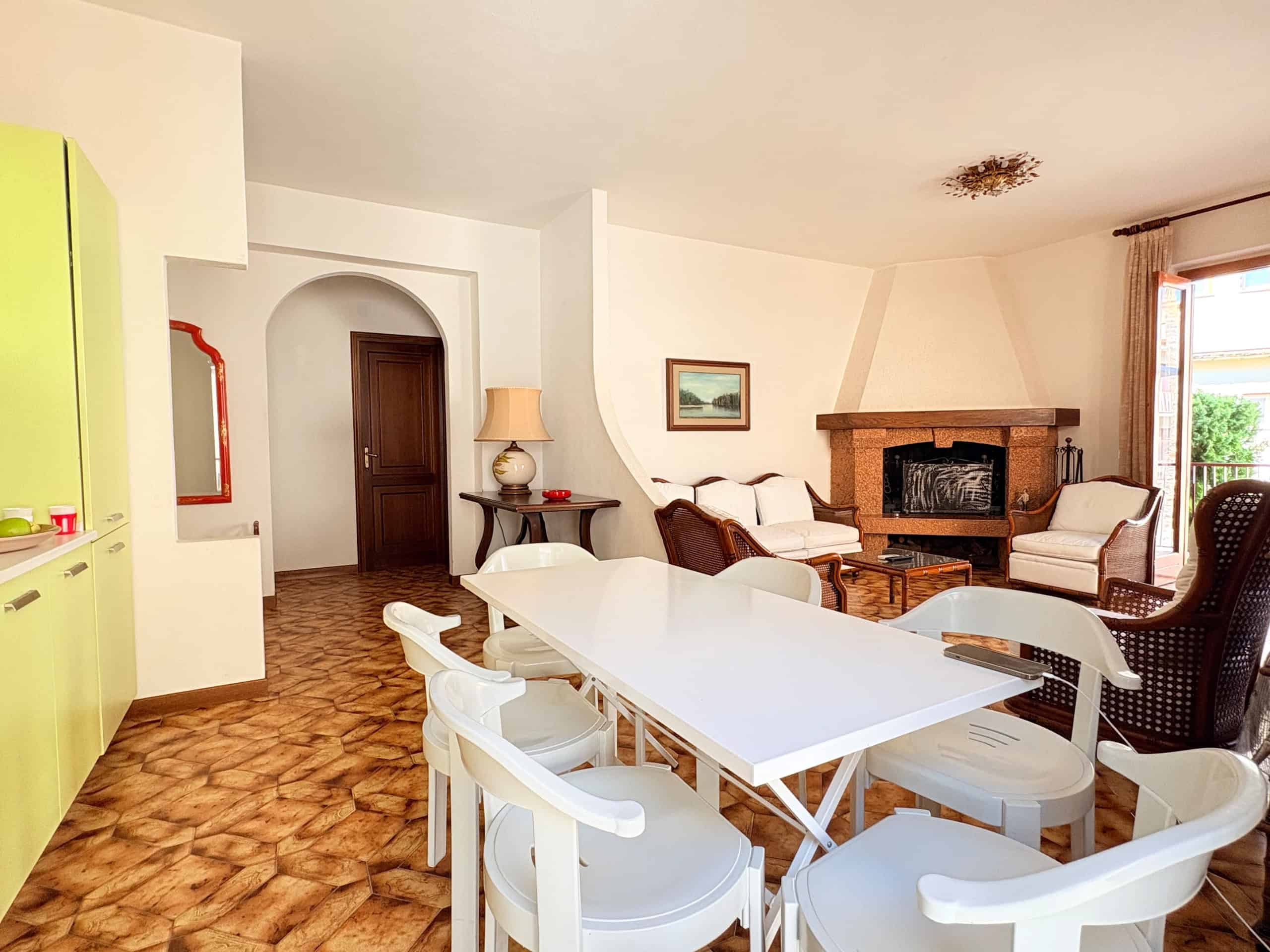 Interno di un soggiorno luminoso con camino in pietra, zona pranzo con sedie bianche e arredi accoglienti, vista su terrazza esterna, stile moderno e confortevole.