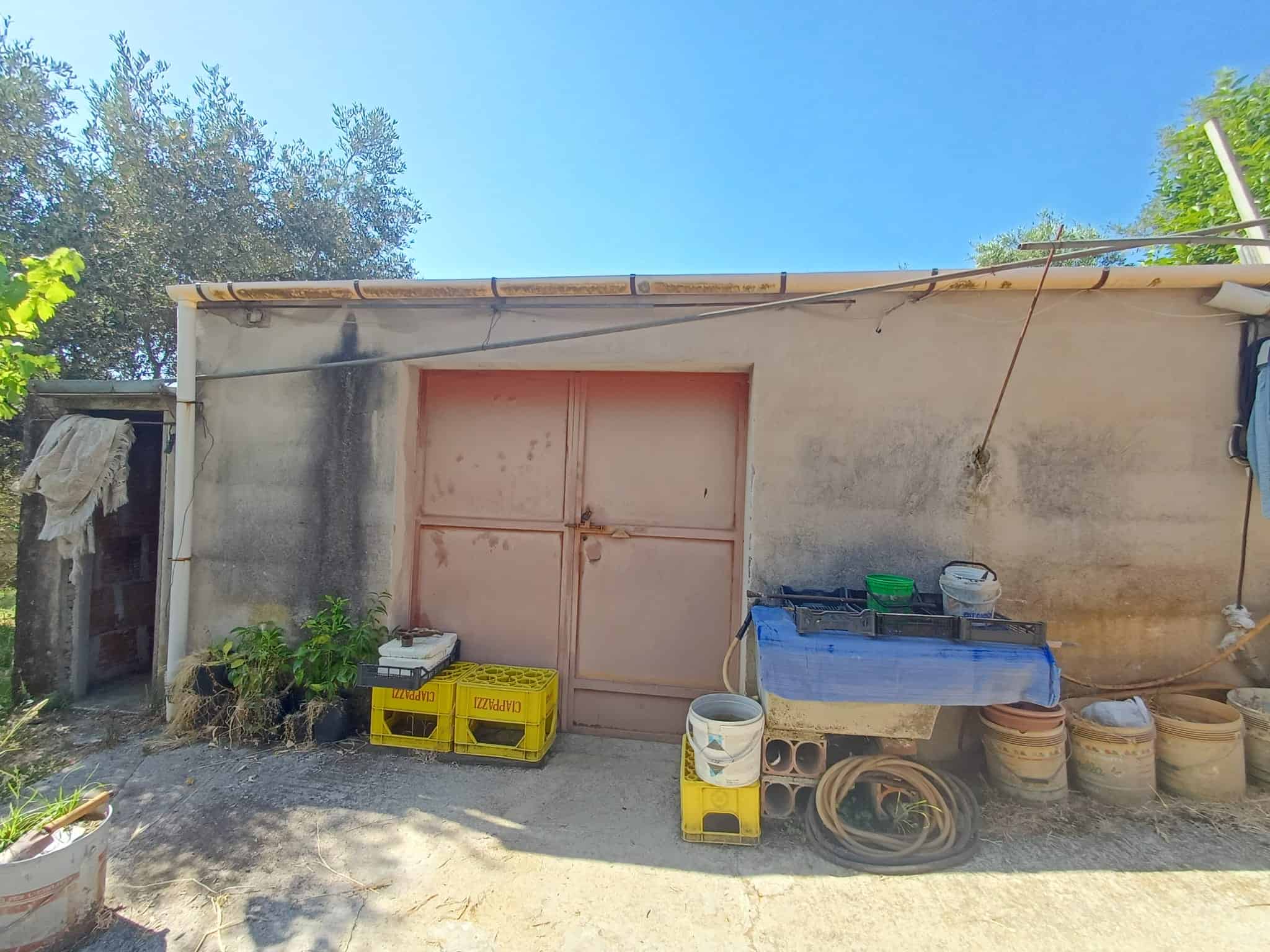 Capannone in muratura con porta in metallo, attrezzato con scatole di plastica, tubi e materiali da giardinaggio, sotto un cielo azzurro, in un'area esterna.
