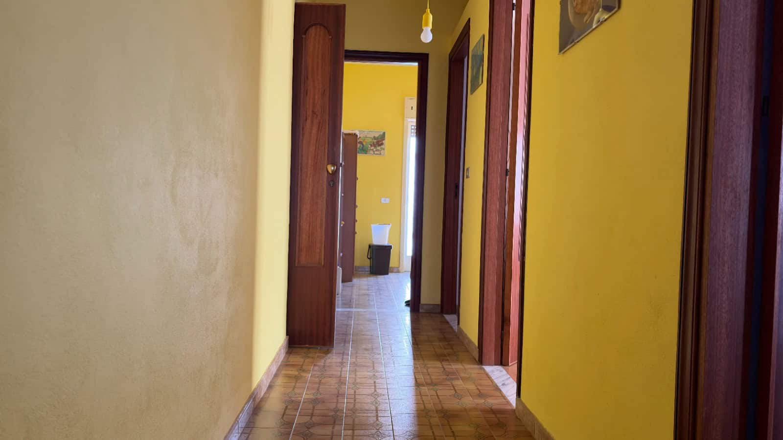 Porte interne in legno e pareti gialle in un corridoio di una casa, vista verso una stanza con pareti bianche e pavimento in piastrelle.