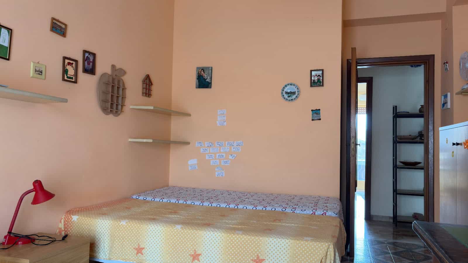 Ampia camera da letto con pareti color pesca, decorata con quadri e oggetti artistici, letto matrimoniale con copriletto a fantasia, lampada da tavolo rossa, e porta aperta che conduce a un'area di se.