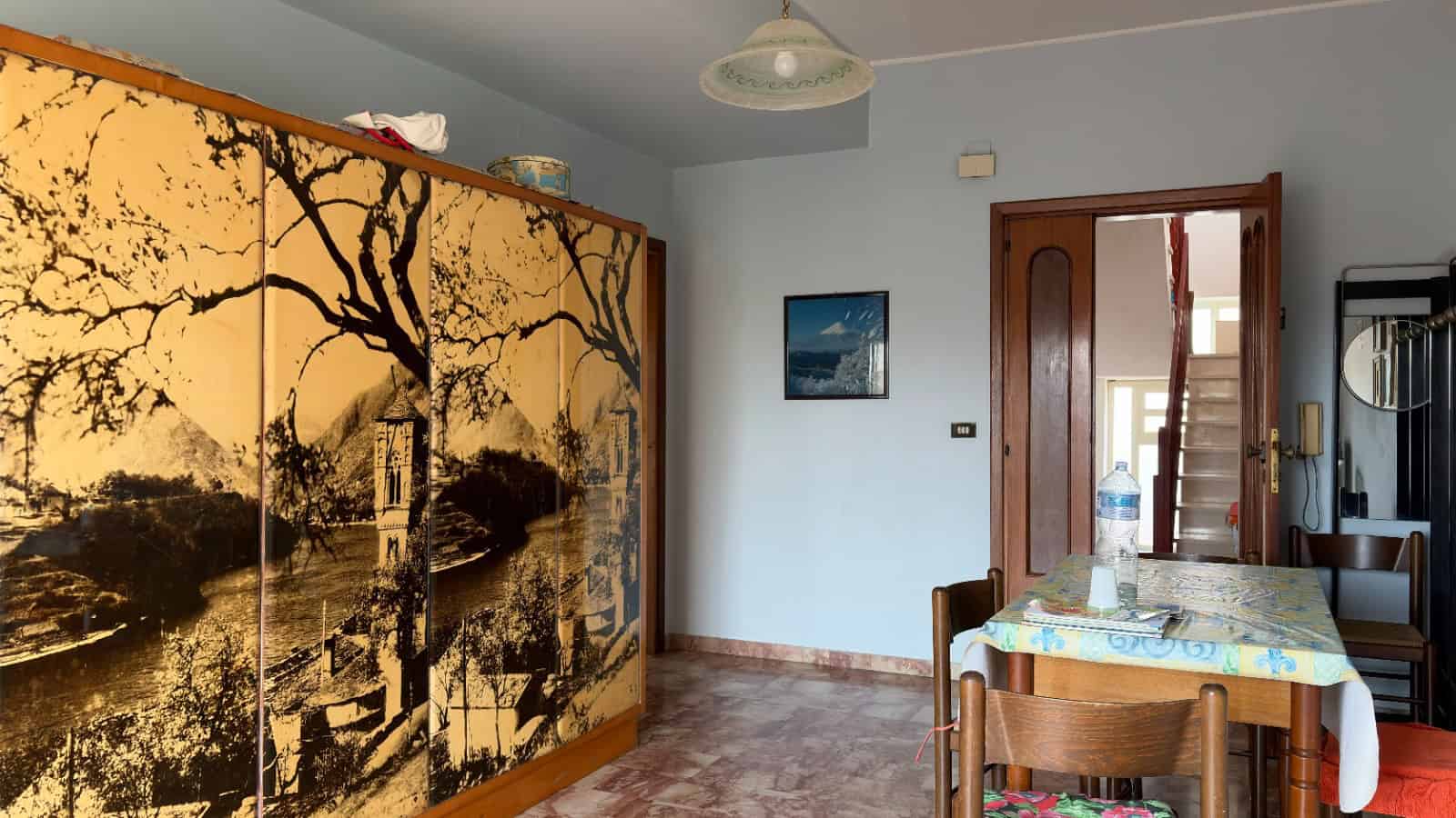 Interno di una casa con armadio a ante decorate con un paesaggio naturale, tavolo con tovaglia e sedie, pareti chiare e porta in legno, ambiente accogliente e luminoso.