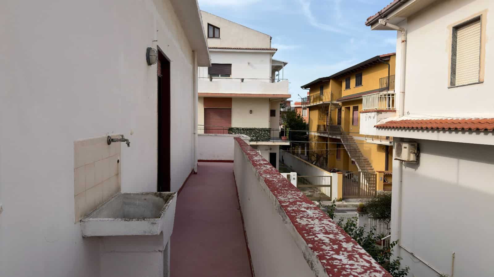 Lavandino esterno su balcone di un edificio residenziale in zona urbana, con case vicine e cielo sereno.