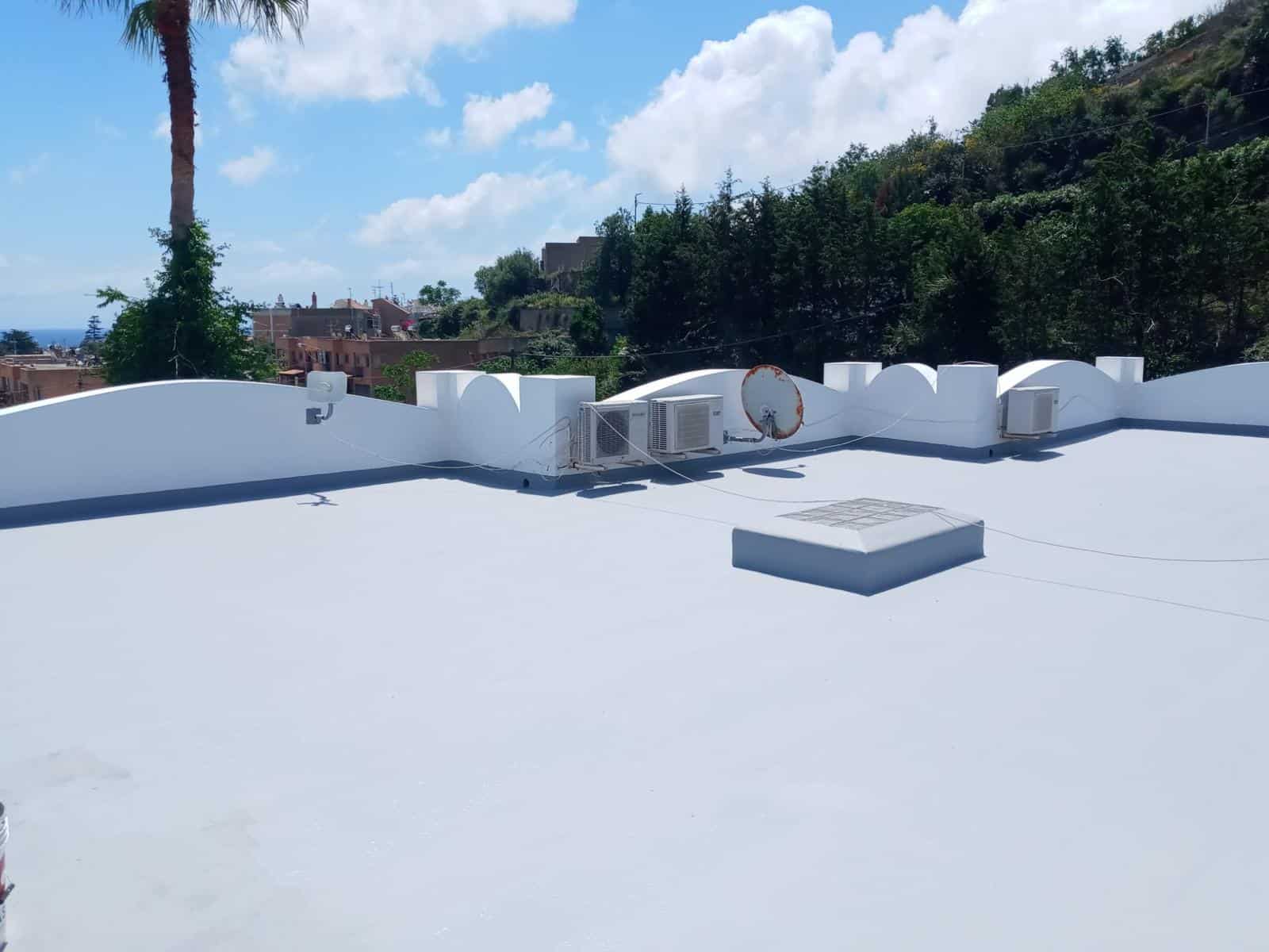 Il tetto bianco di un edificio con vista su un paesaggio verde e cielo sereno, con elementi di ventilazione e condizionamento, in un'area residenziale soleggiata.