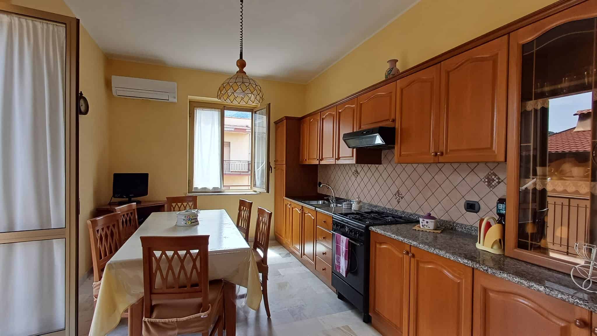 Pannello di cucina in legno con piano in granito, zona pranzo con tavolo e sedie, finestra luminosa, elettrodomestici moderni, ambiente accogliente e funzionale.