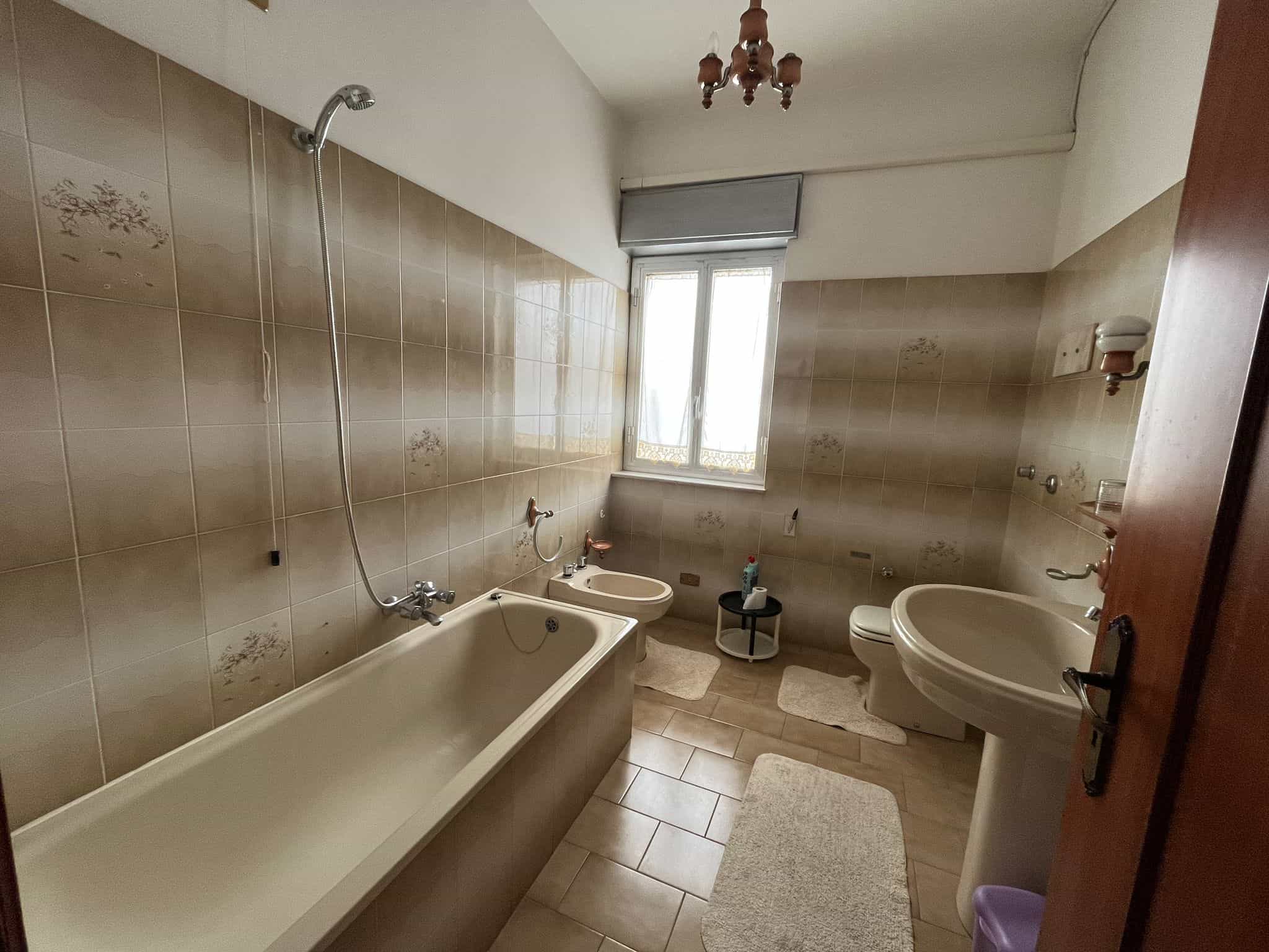 Acqua calda e ambiente luminoso in bagno con vasca, lavabo e WC, finestra con vista, piastrelle beige, arredo semplice, ideale per ristrutturazione o investimento immobiliare.
