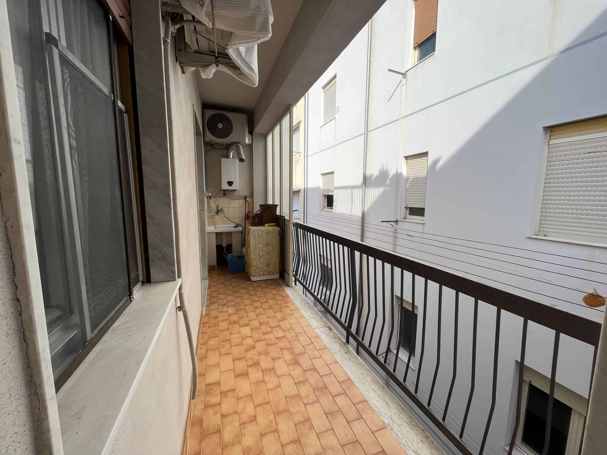 Balcone di un appartamento con pavimento in piastrelle, ringhiera in metallo e vista su edifici circostanti, in un contesto urbano residenziale.