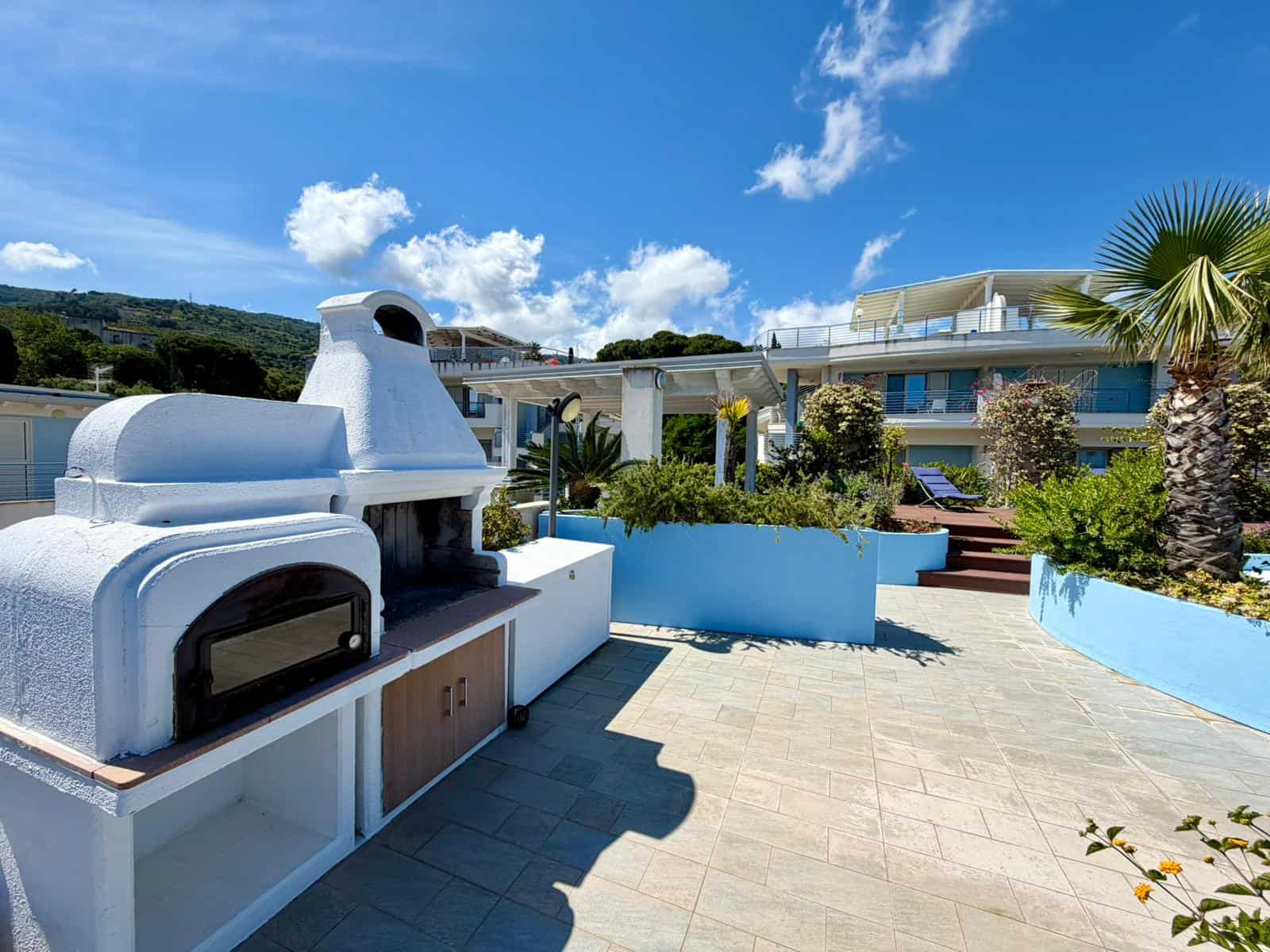 Forno a legna esterno con vista panoramica su terrazza soleggiata e giardino ben curato, ideale per cene all'aperto e relax in famiglia. Design moderno e funzionale, perfetto per case di lusso.