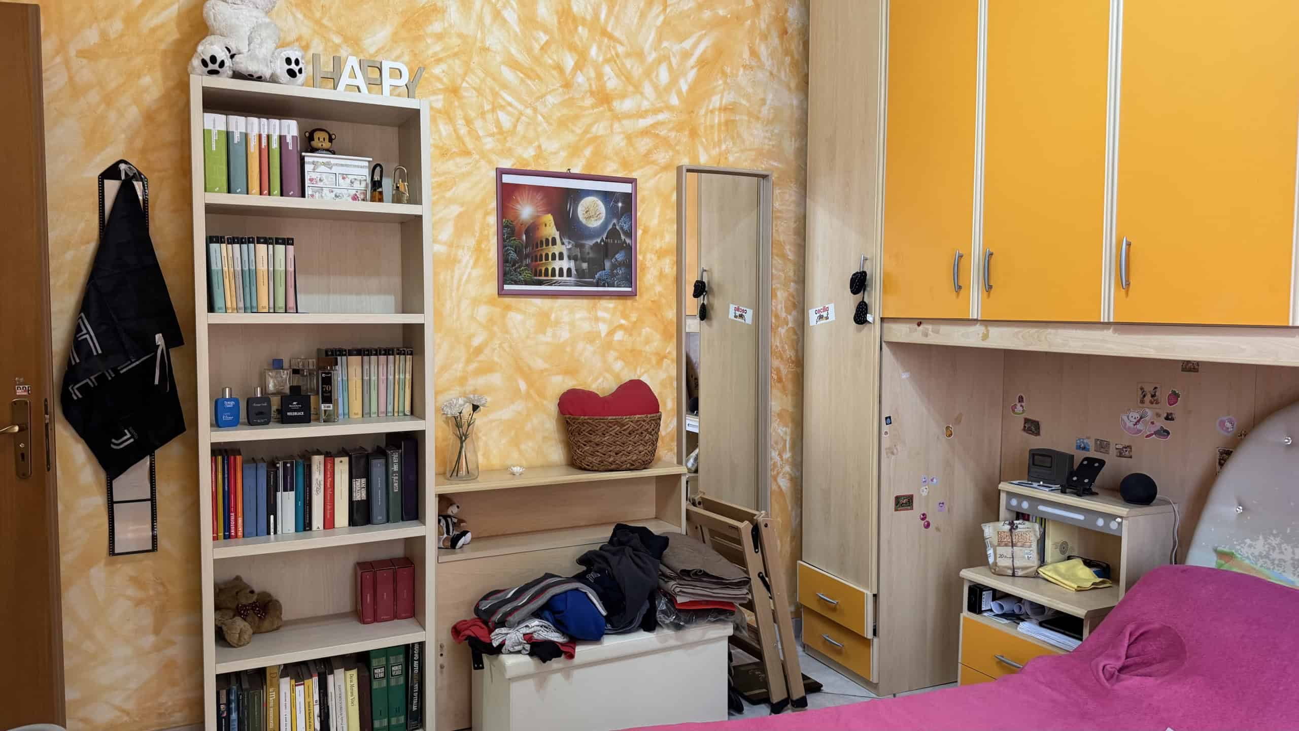 Spazio di una camera da letto con armadio giallo, scaffale con libri, letto con vestiti e decorazioni colorate, ambiente accogliente e vivace.