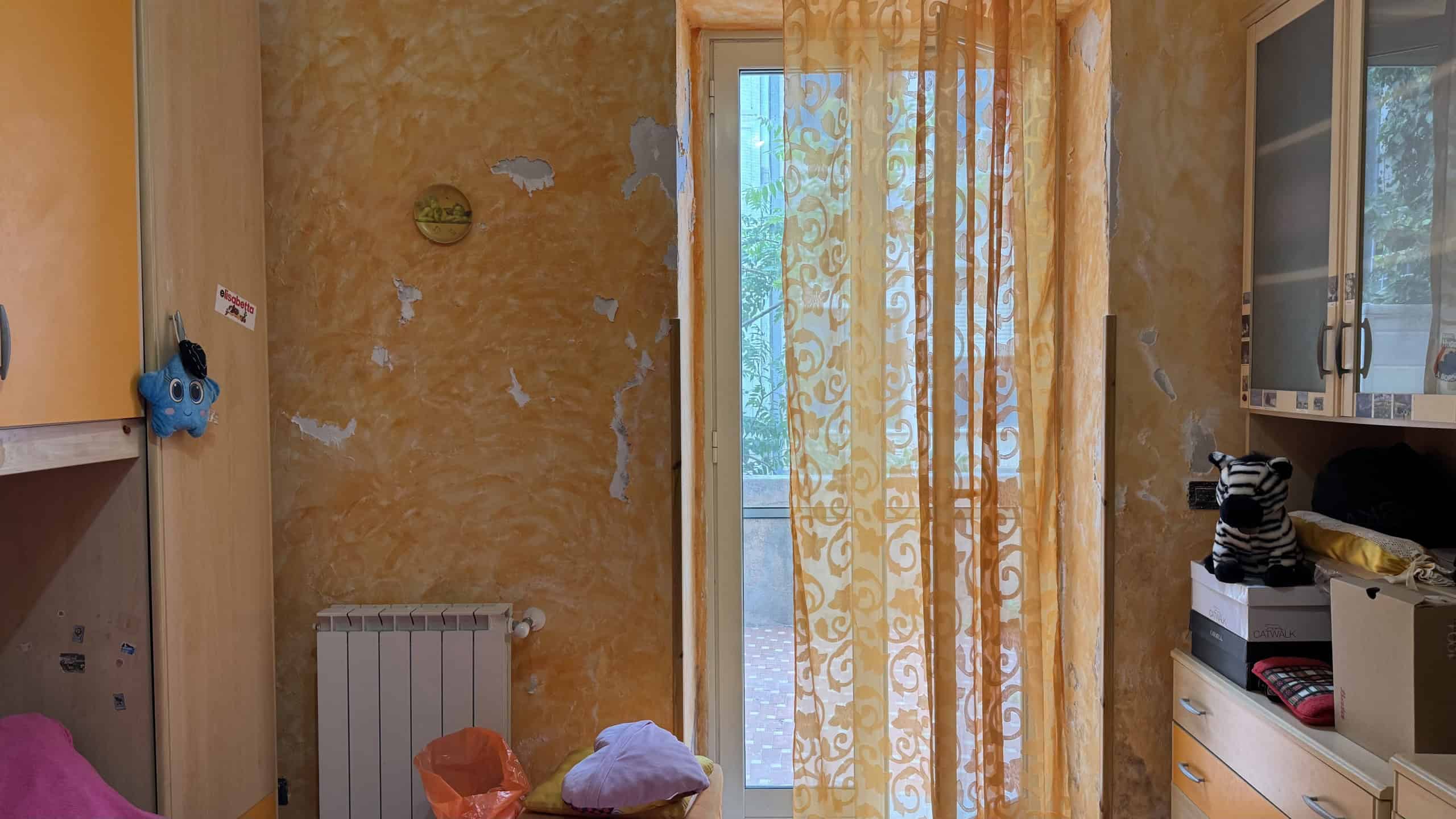 Soggiorno con pareti gialle, porta finestra con tende decorative che conduce a un balcone, arredamento semplice e funzionale, ambiente abitabile e luminoso.