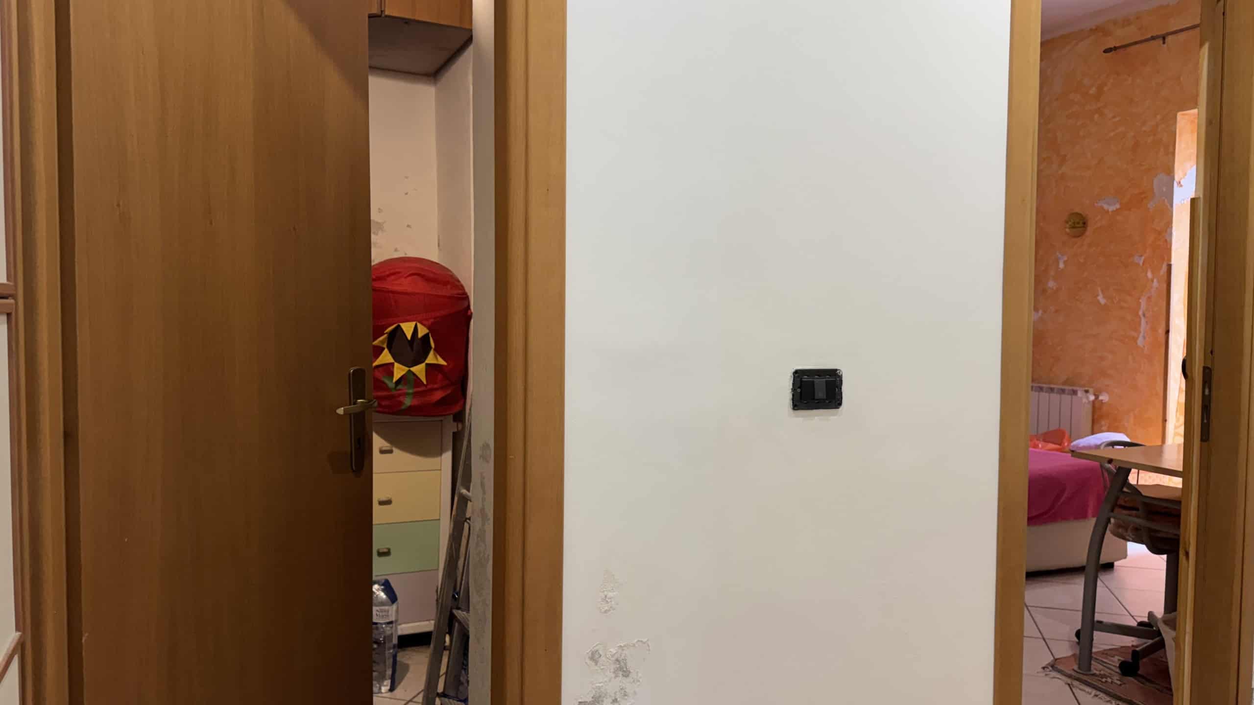Spazio interno di una casa con pareti bianche, porta in legno aperta e dettagli di arredamento visibili, come un armadio e una sedia, in un ambiente domestico accogliente e luminoso.