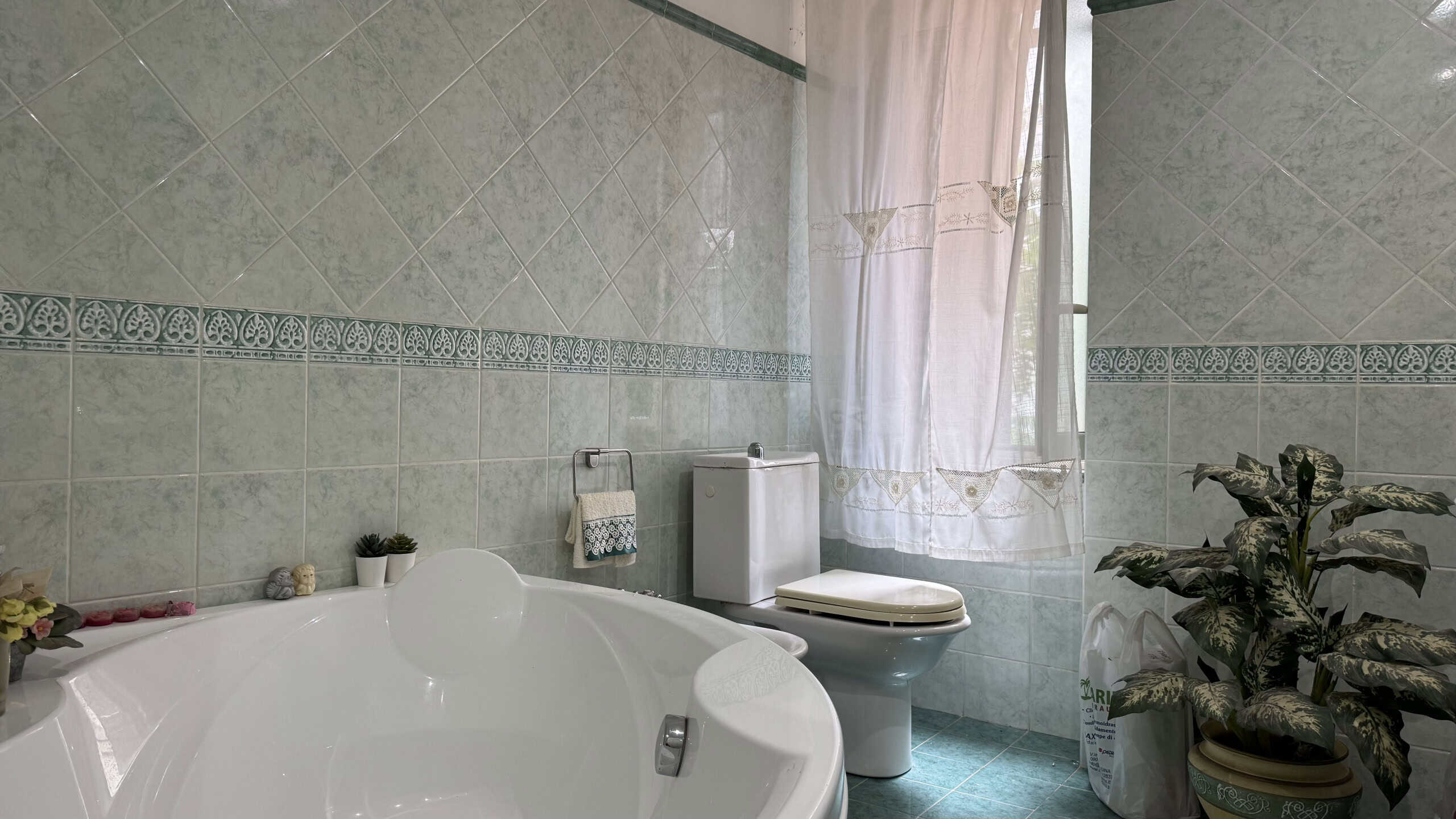 Lavabo e bagno con piastrelle decorative, finestra con tende leggere, piante verdi e arredi moderni in un ambiente luminoso e accogliente.