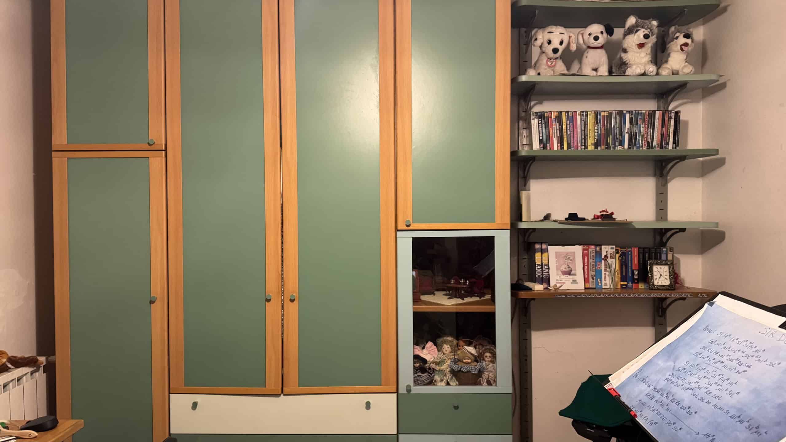 Spazio di soggiorno con armadio in legno e pareti verdi, decorato con bambole e libri su scaffali, creando un ambiente accogliente e familiare.