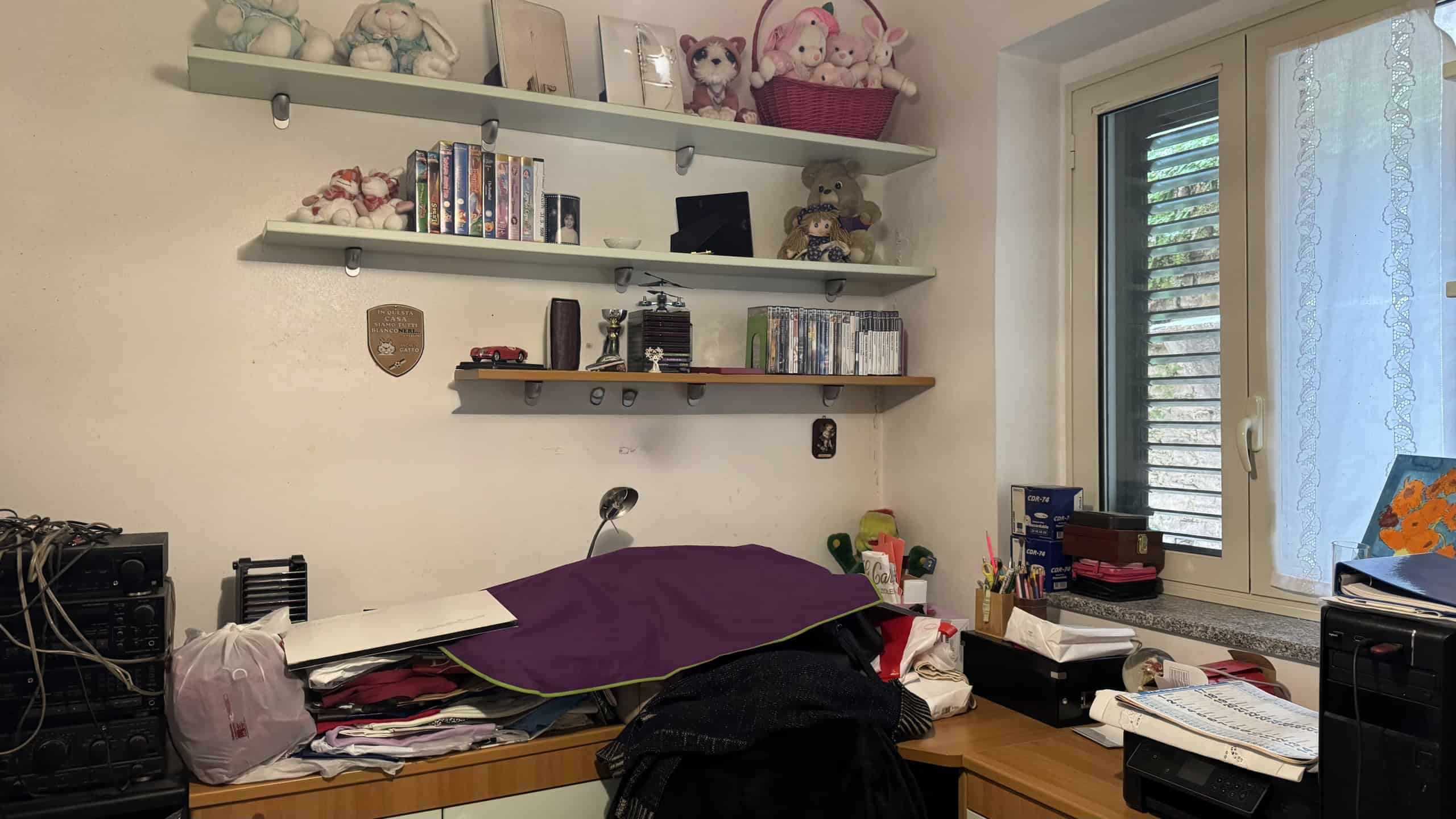 Spazio di lavoro domestico con scrivania, libri, oggetti personali e decorazioni, illuminato da finestra con persiane. Ambiente ordinato e funzionale per studio o attività creative.