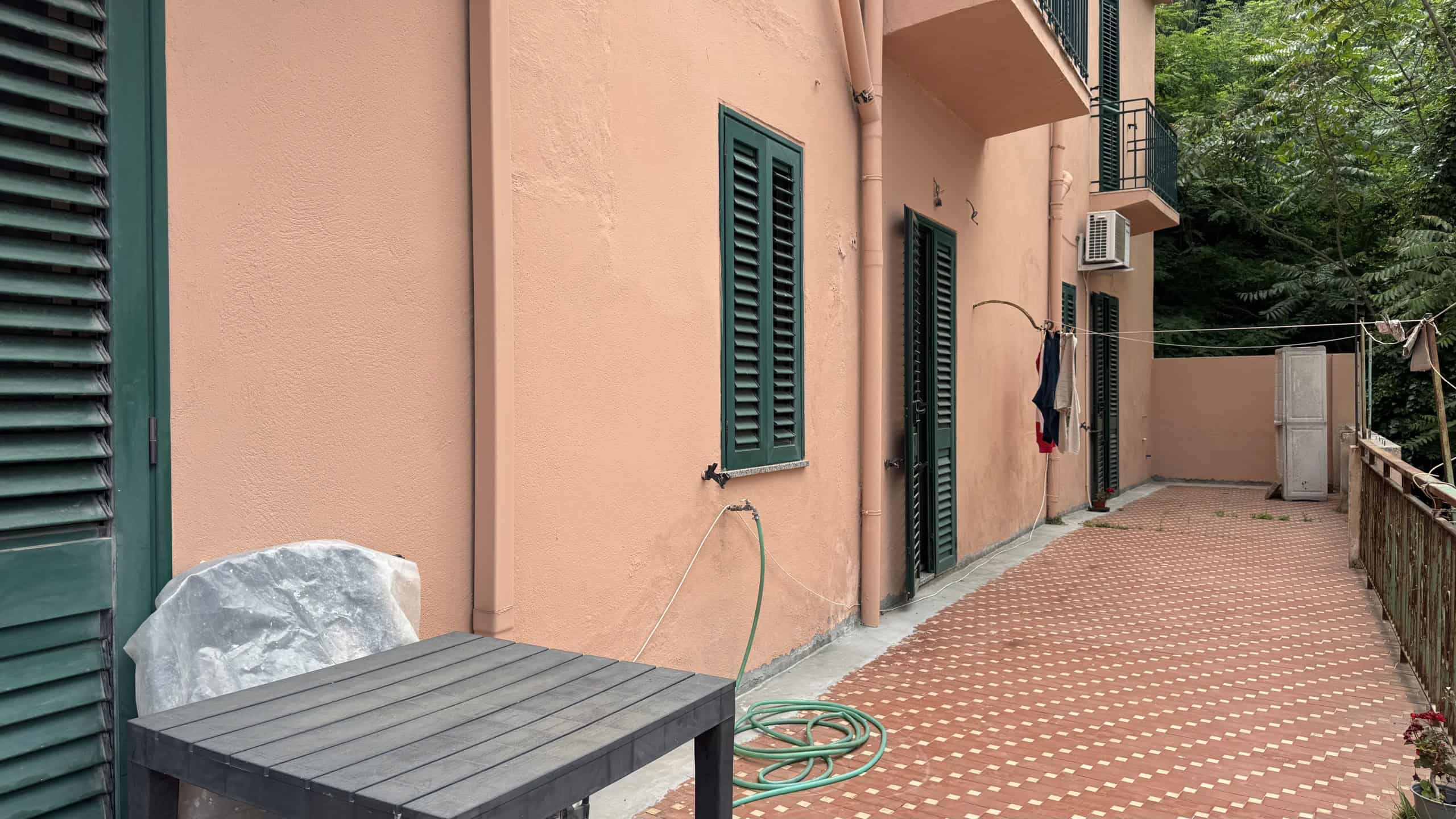 Spiaggia di un appartamento con pareti color pesca, finestre con persiane verdi e un balcone, con un cortile esterno pavimentato e un'area verde sullo sfondo, in un ambiente residenziale tranquillo.