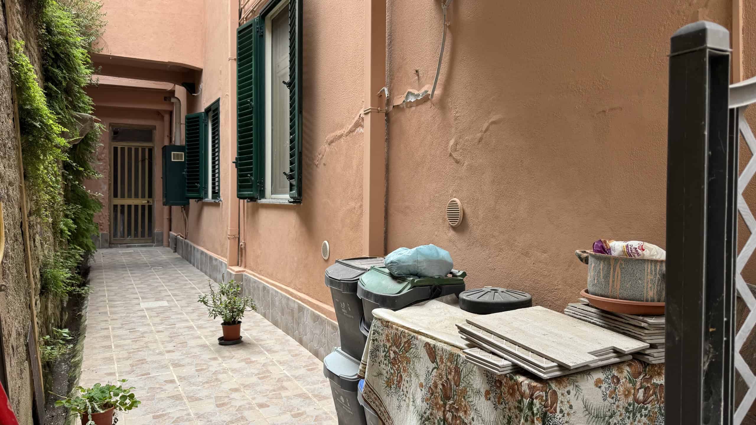 Spazzatura e materiali di riciclo accatastati vicino a una parete di un edificio con finestre con persiane verdi, in un cortile urbano.