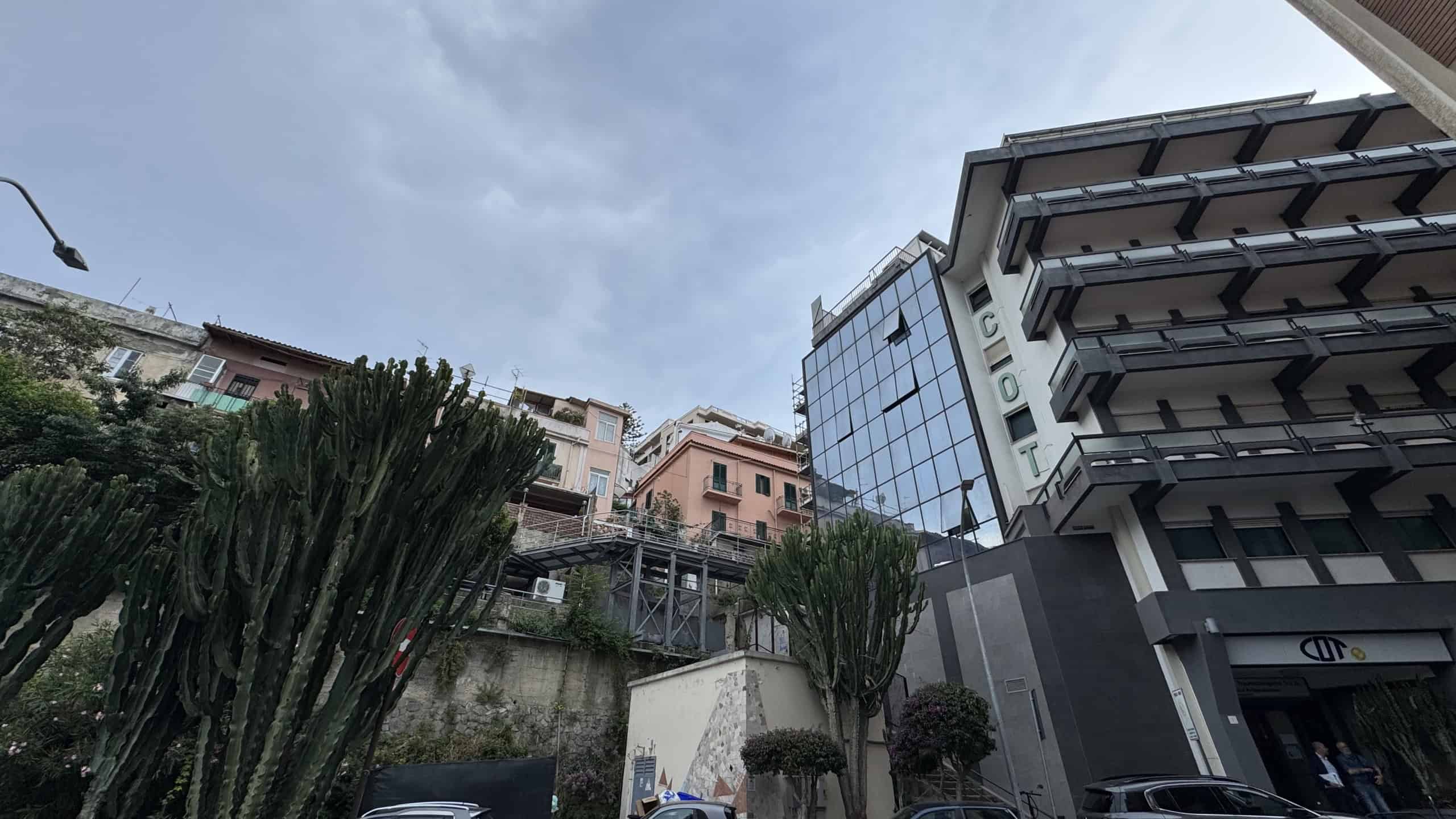 Elegante edificio moderno con facciata in vetro e balconi, situato in un'area urbana collinare, circondato da vegetazione e strutture residenziali, rappresenta un esempio di architettura contemporanea.