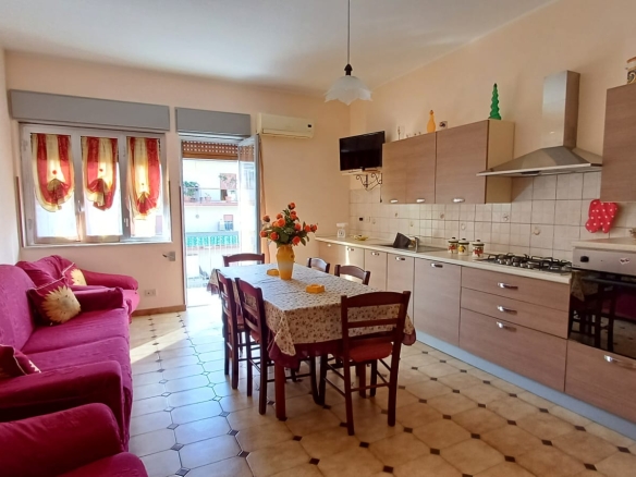 Cucina moderna con soggiorno spazioso, mobili in legno chiaro, tavolo da pranzo con sedie, divano rosso e decorazioni colorate, ambiente luminoso e accogliente.