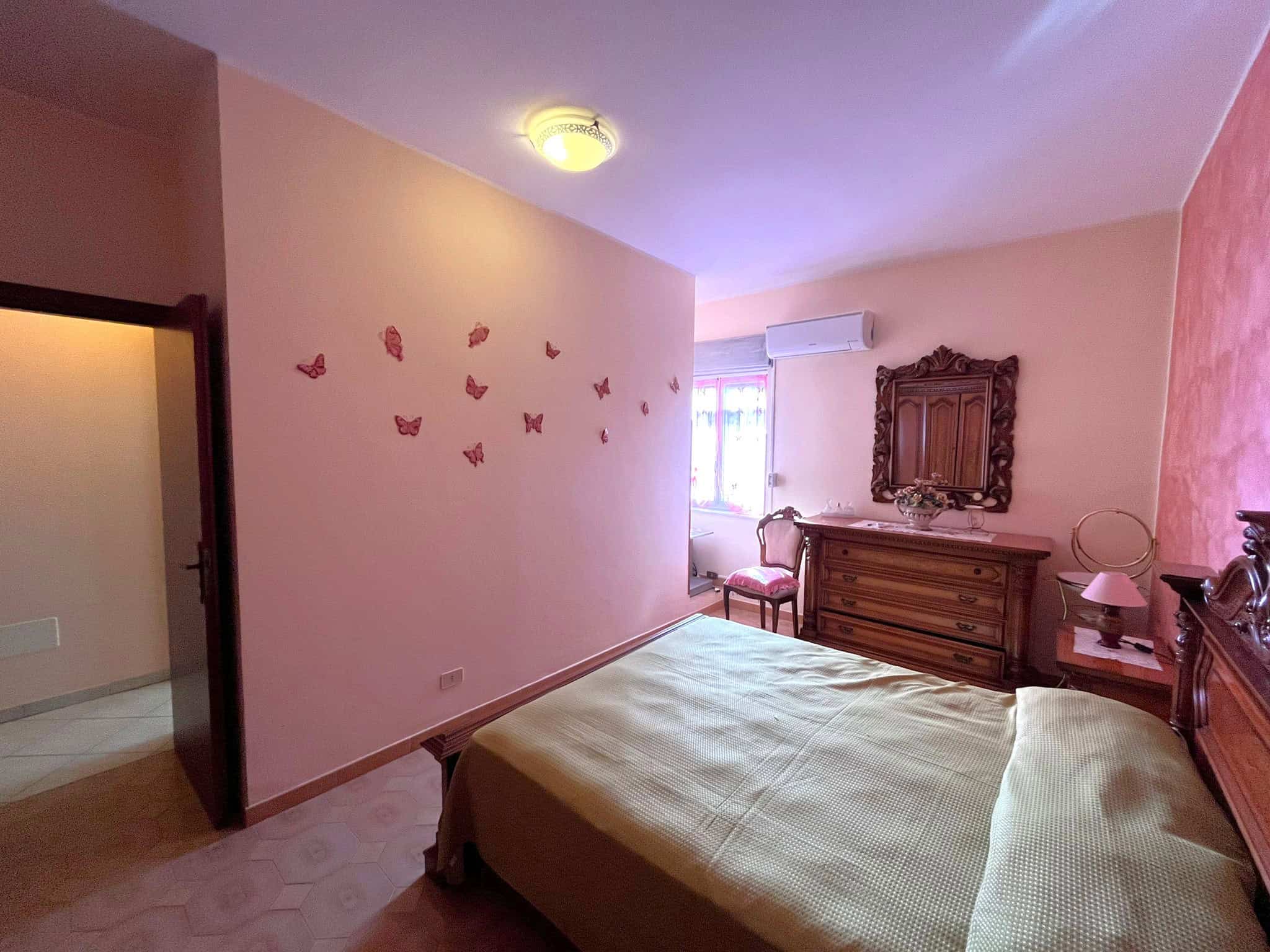 Camera da letto spaziosa con pareti rosa, decorazioni di farfalle e mobili in stile classico, illuminata da luce naturale e lampada a soffitto, ambiente accogliente e elegante.