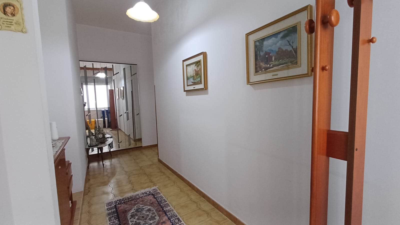 Ampia vista di un corridoio interno di una casa con pareti bianche, decorato con quadri e un tappeto, che conduce a una zona soggiorno luminosa e accogliente.